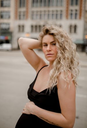 Woman in black dress with flowing blonde curls poses against urban backdrop.