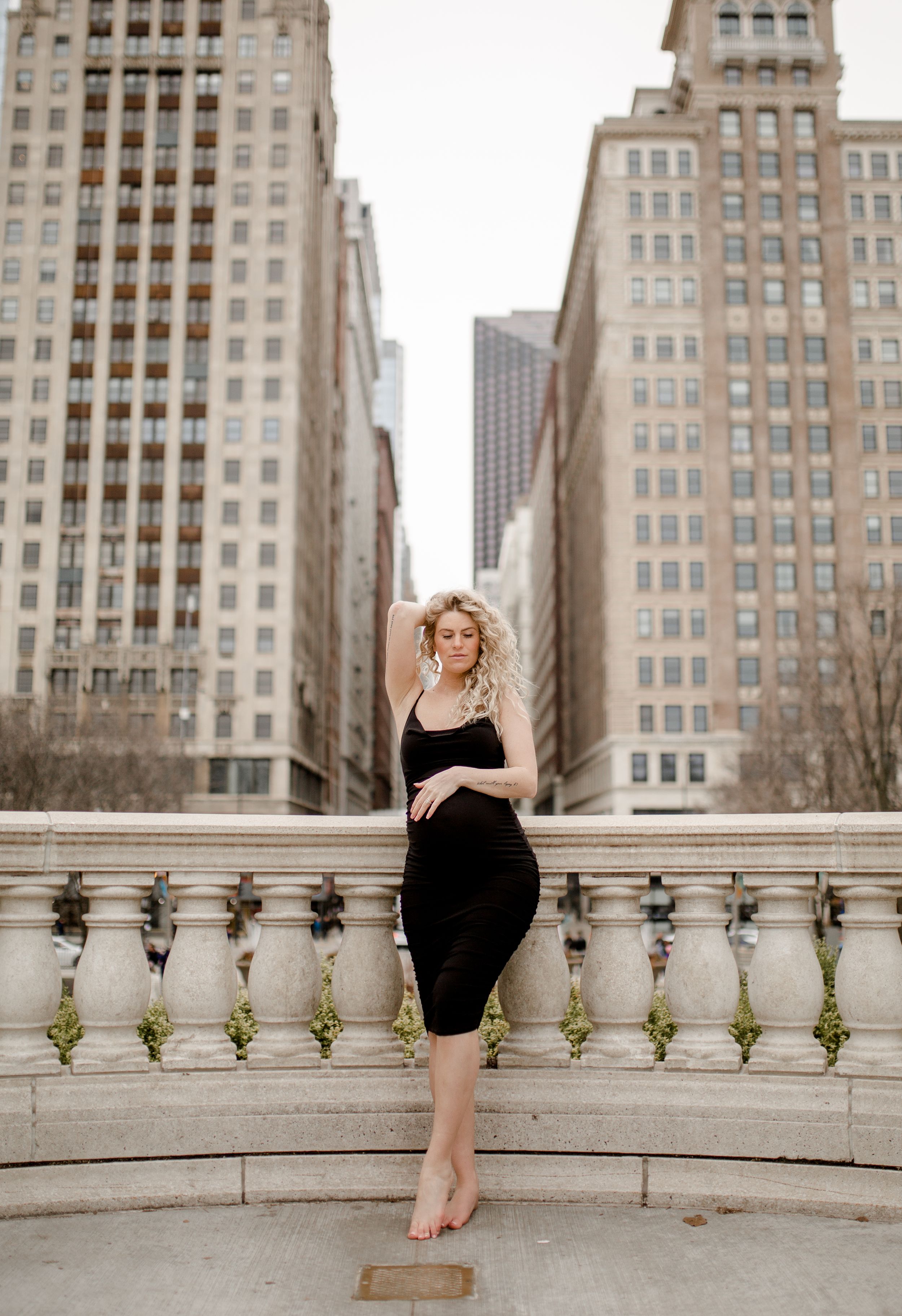 Woman in black dress poses on stone balustrade with tall city buildings behind her.