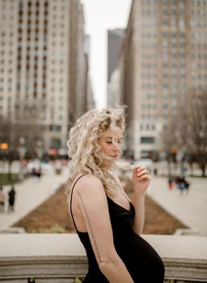 Woman in black dress poses on stone ledge with urban plaza and skyscrapers visible.