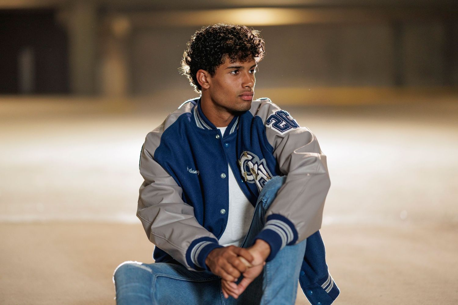 A person wearing a blue and gray varsity jacket sits thoughtfully in a dimly lit setting.