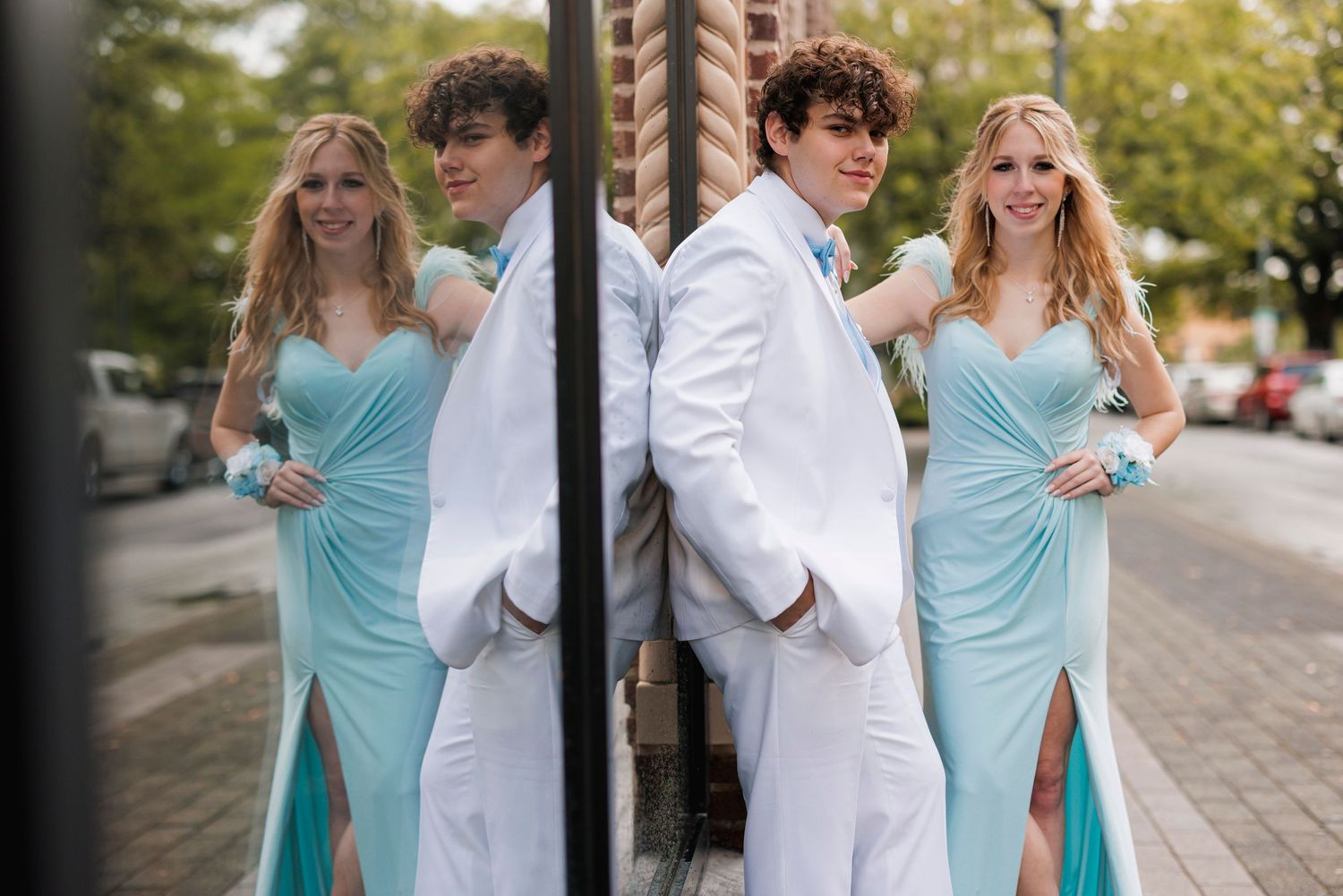 Two couples in matching aqua dresses and white suits pose elegantly against a brick pillar on a city street.