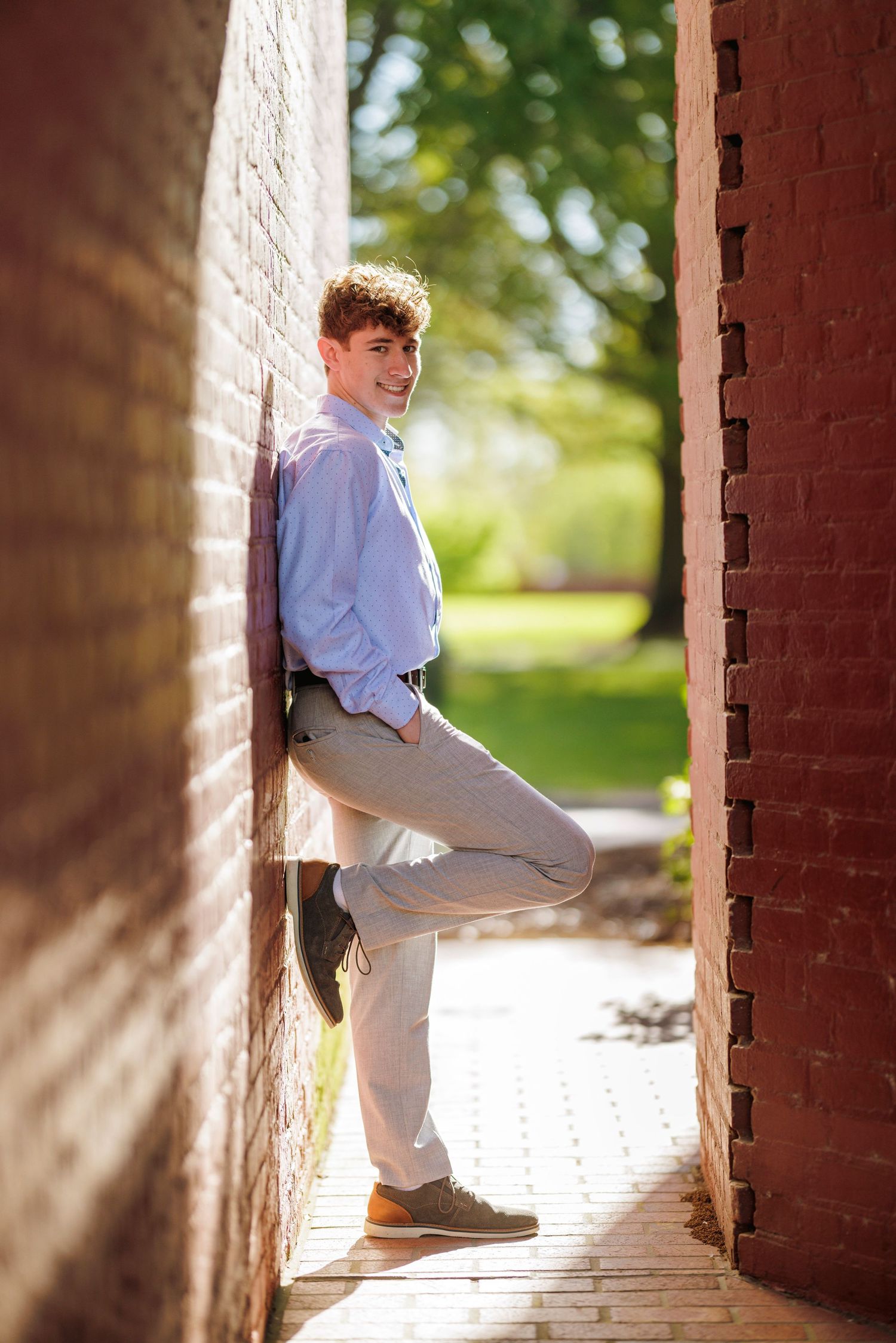 A person in light business casual attire poses against a brick wall in natural sunlight.