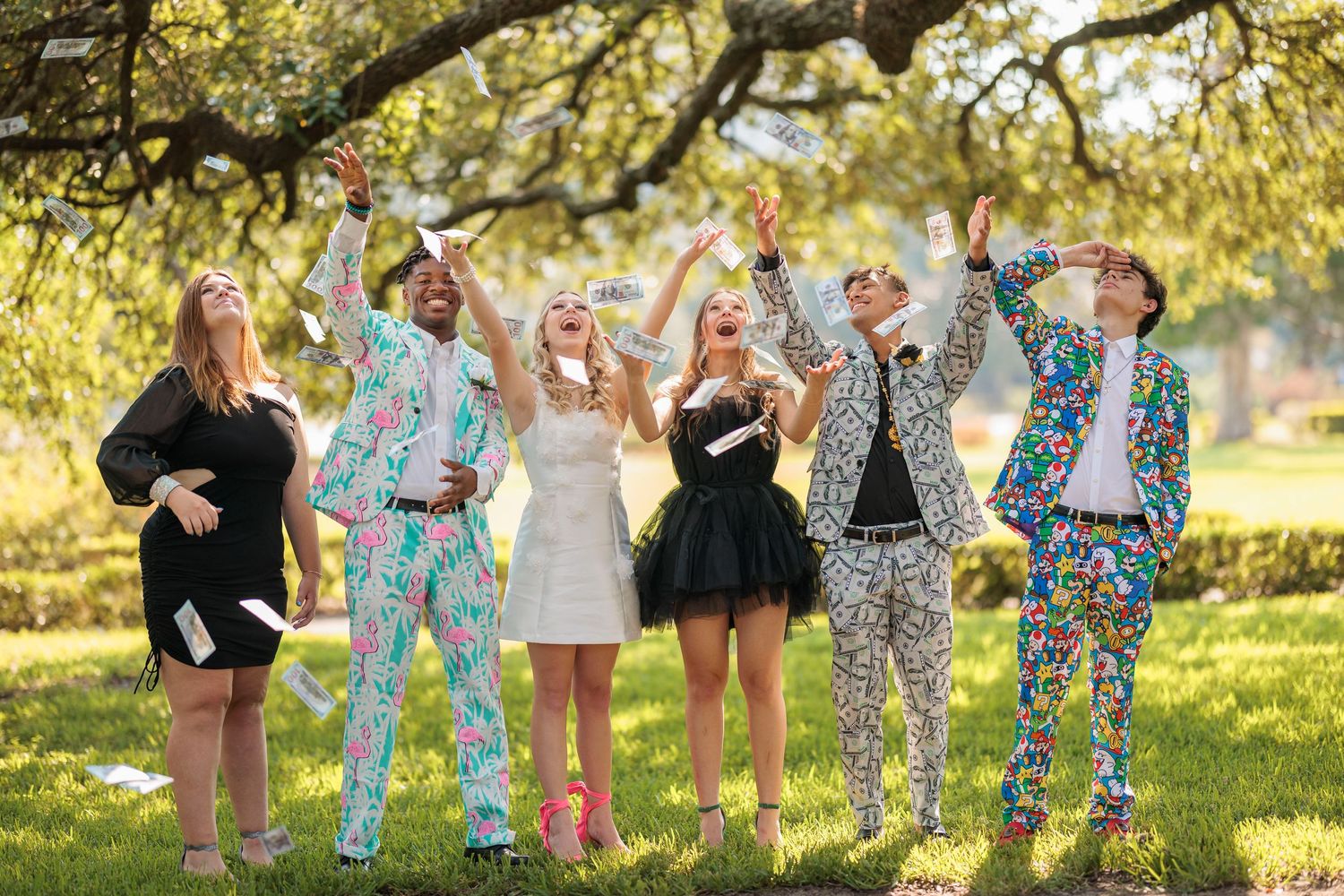 A group poses joyfully outdoors wearing colorful formal attire under trees with golden sunlight.