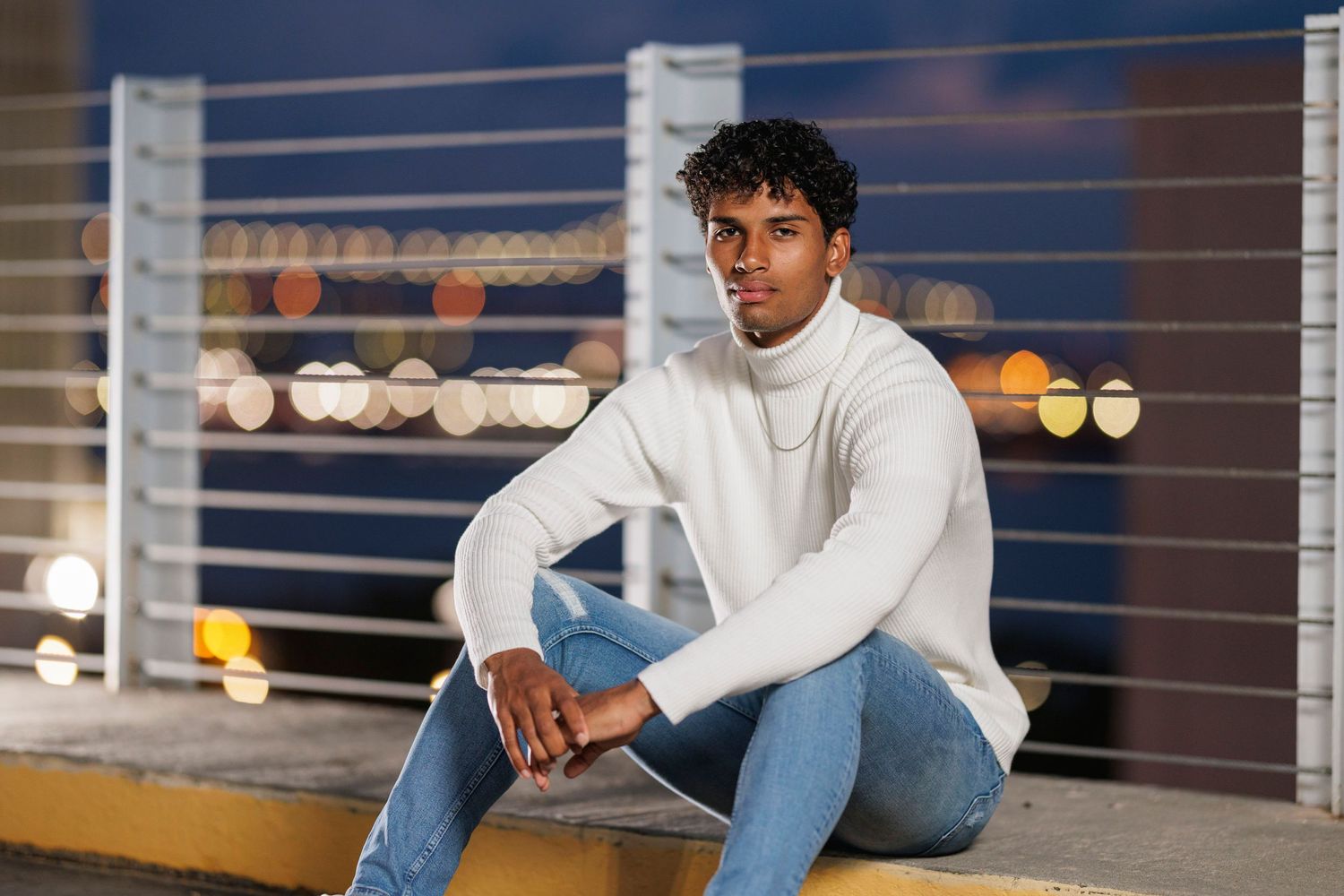 A person in a white turtleneck sweater and jeans sits casually on an outdoor ledge at night.