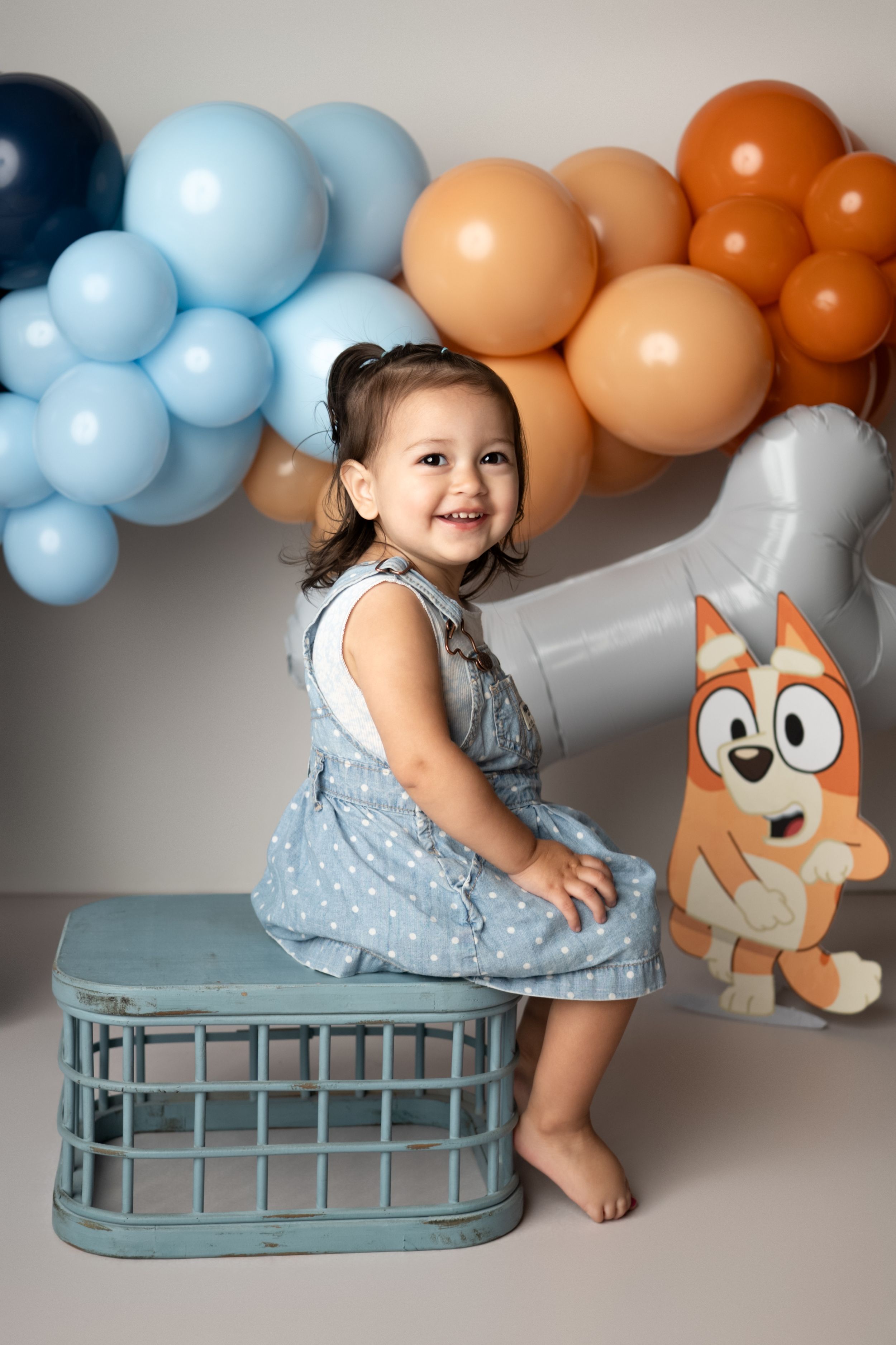 Baby in silver dress sits on blue bench with orange and blue balloon garland and fox plush toy decorations.