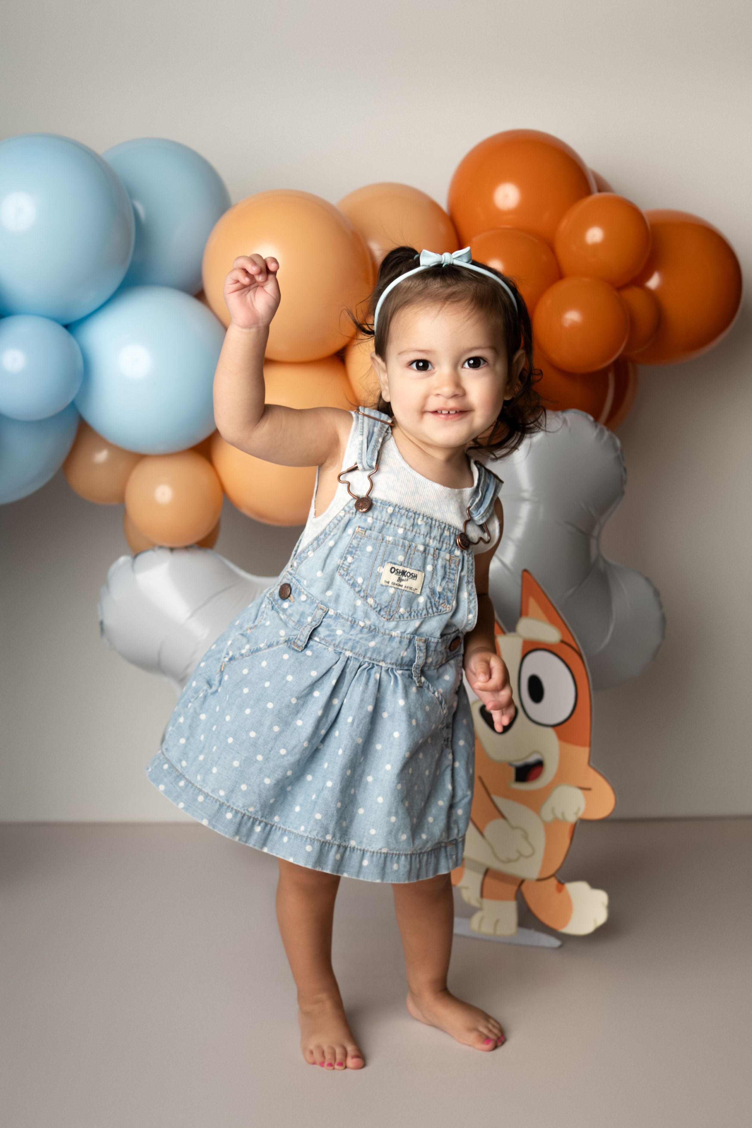 Small child in blue polka dot dress poses playfully against wall decorated with orange and blue balloons.