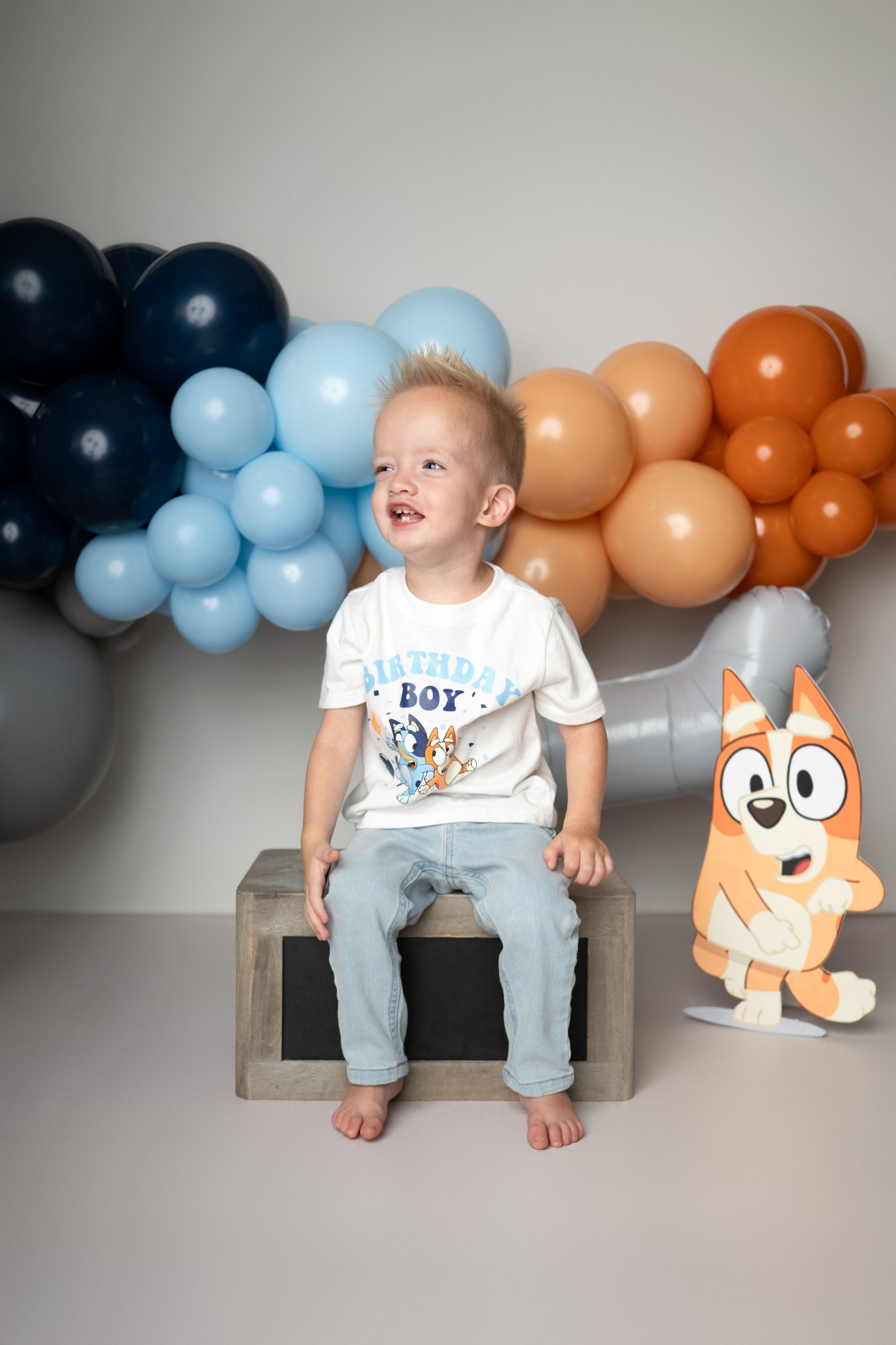 A child sits on a wooden bench with blue and orange balloons and a plush toy against a gray background.