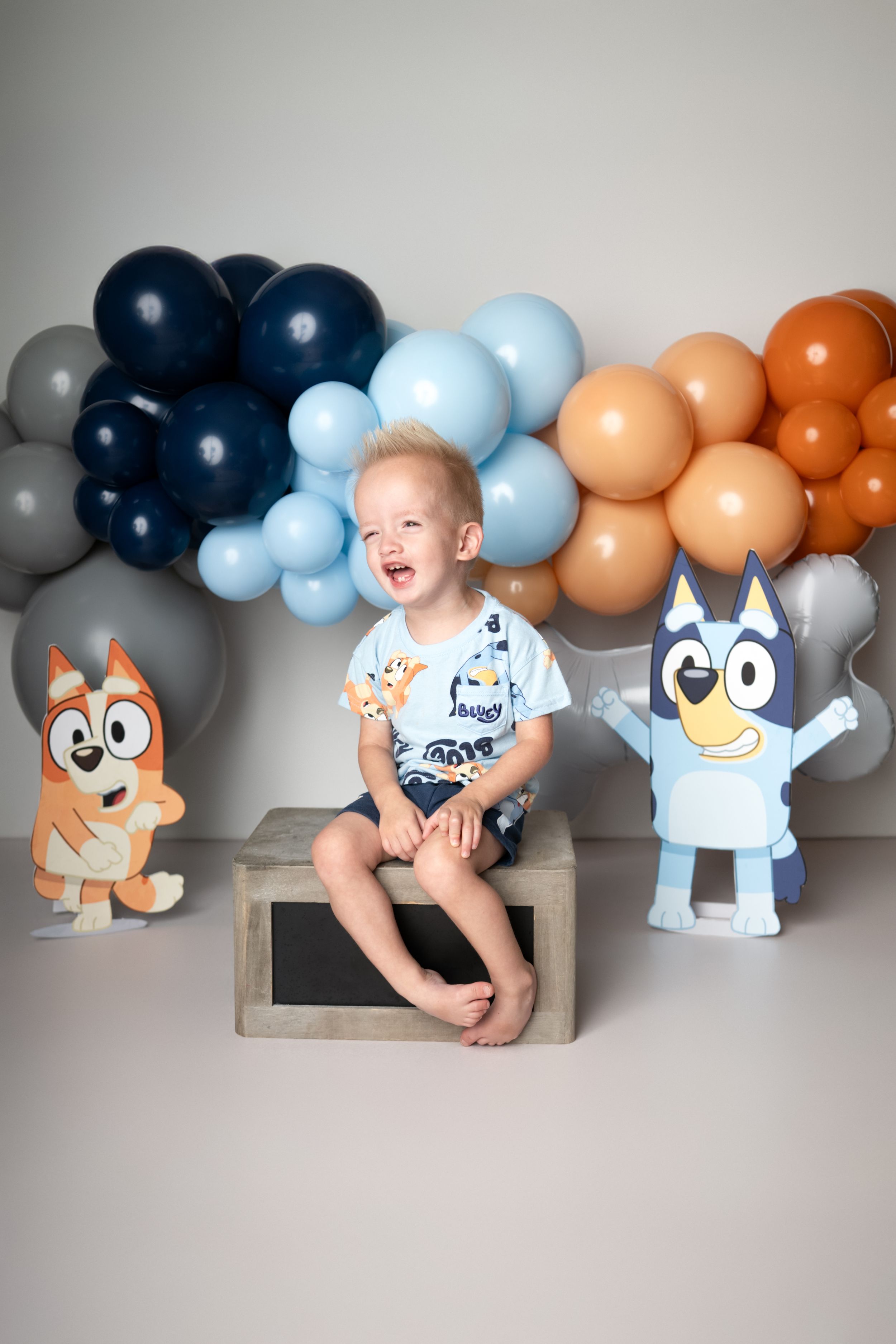A baby's monthly milestone photo series with blue and orange balloon garland and cartoon dog cutouts in the background.