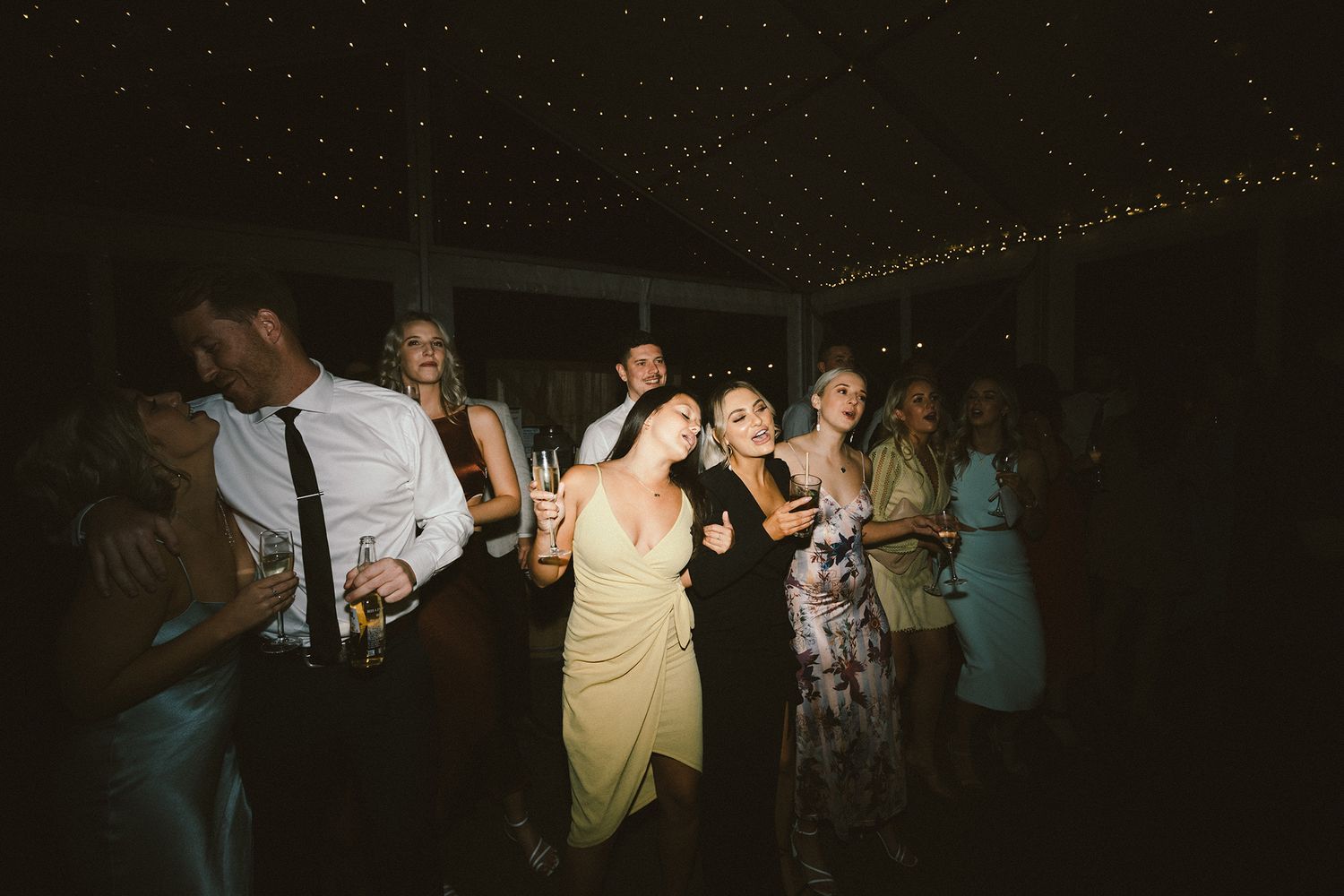 People dancing and celebrating at a dimly lit wedding reception with string lights above.