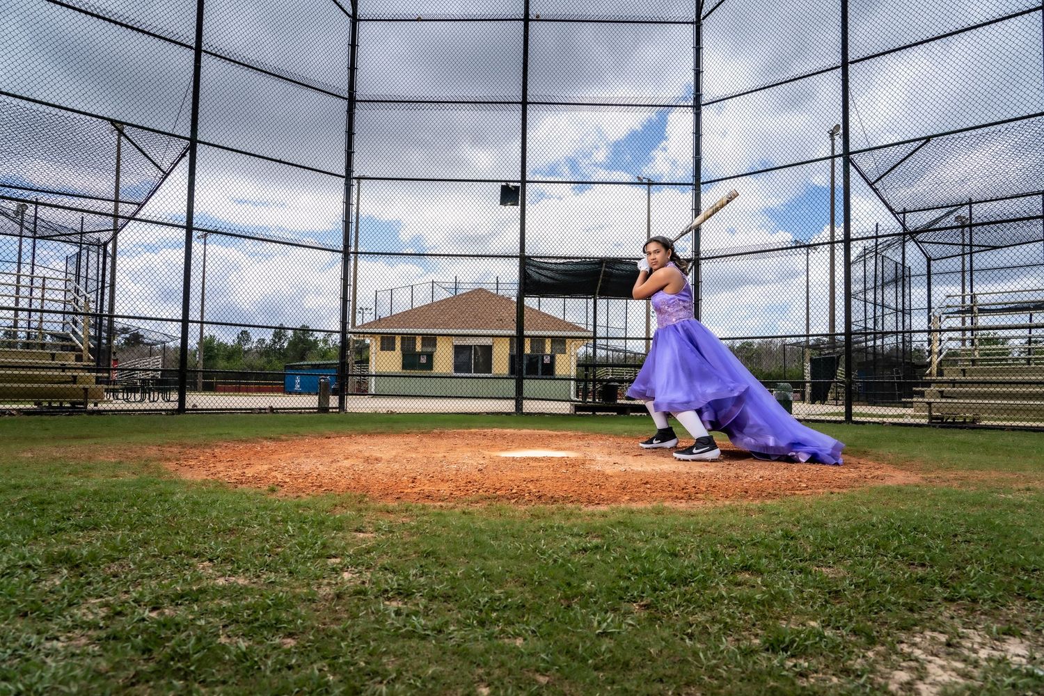 Purple formal dress contrasts with baseball field setting under cloudy skies.