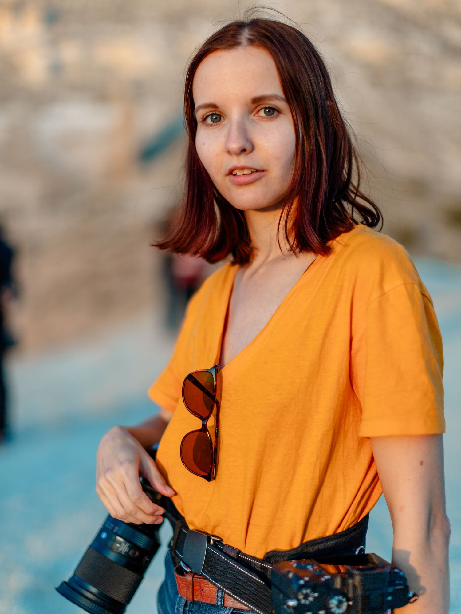 Portrait of photographer wearing bright orange shirt with cameras against blurred background.