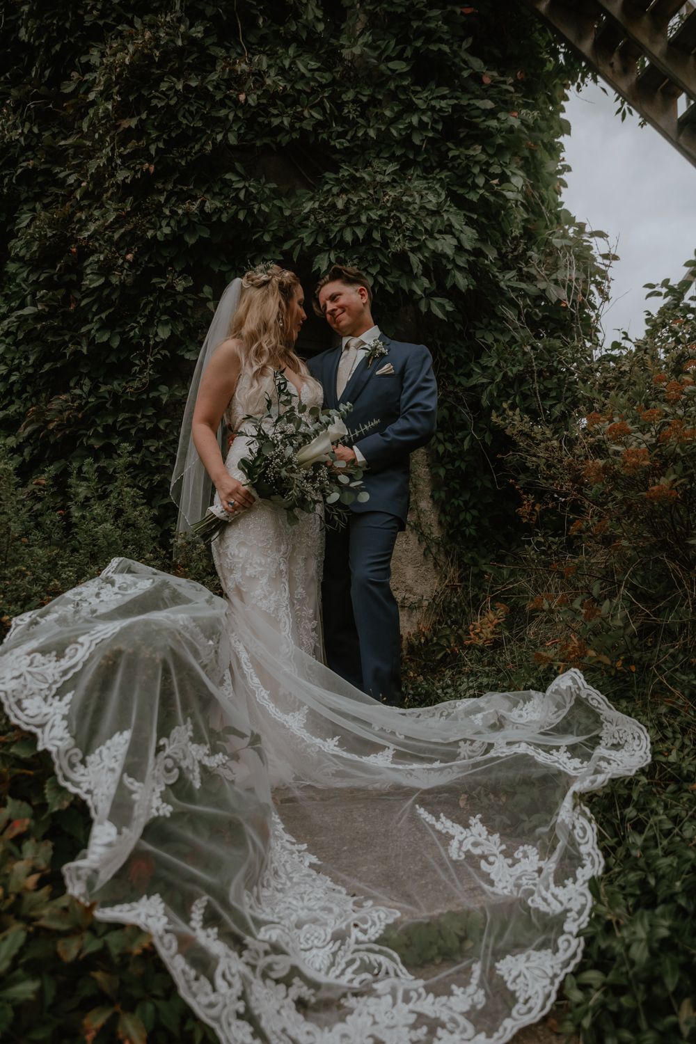a bride and groom gaze at each other in front of an ivy covered tower as the veil cascades in front of them