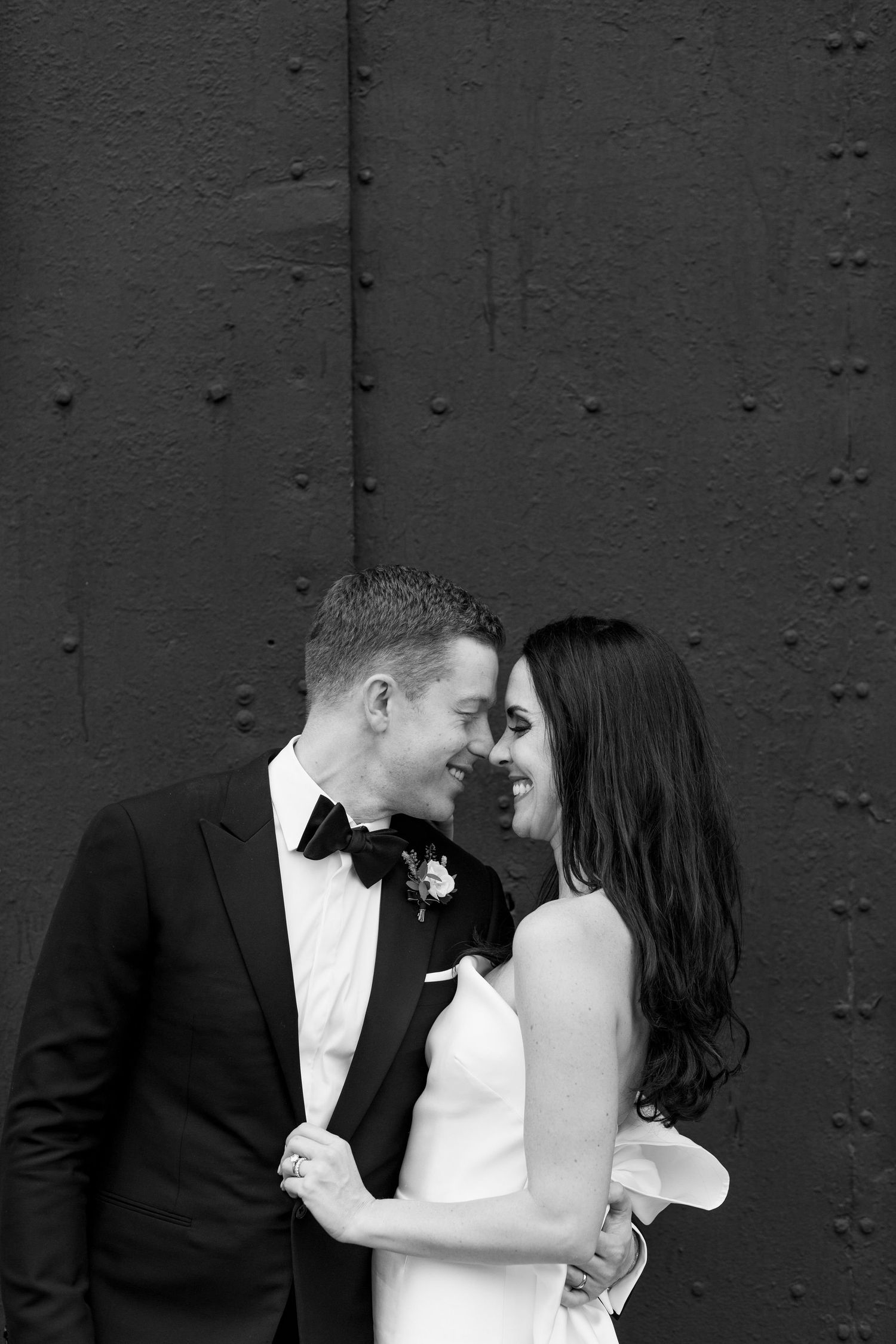 A romantic black and white portrait of a couple in formal wedding attire against a dark textured wall.