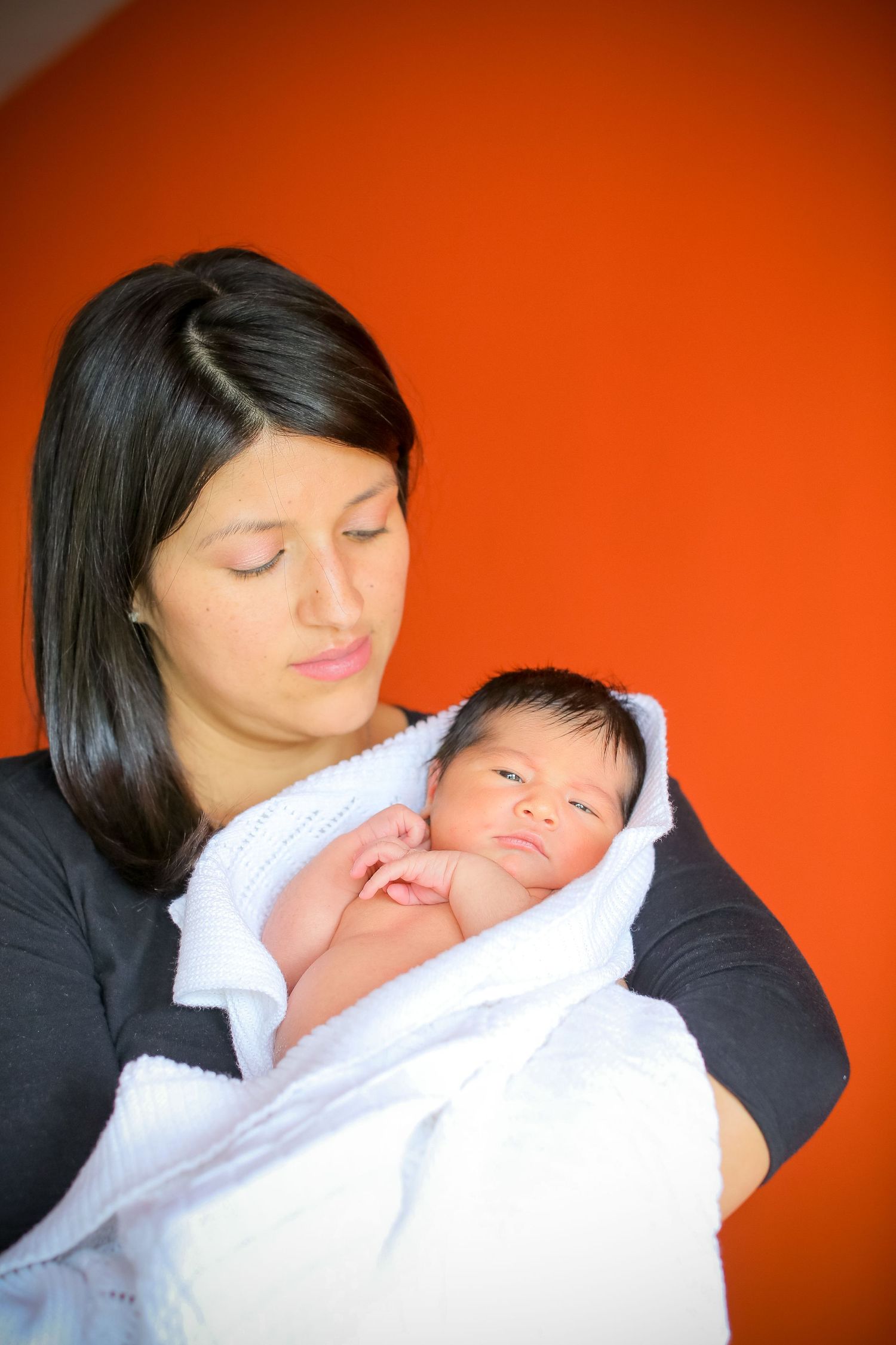 Tender moment of a baby being cradled in white blankets against a vibrant orange wall.