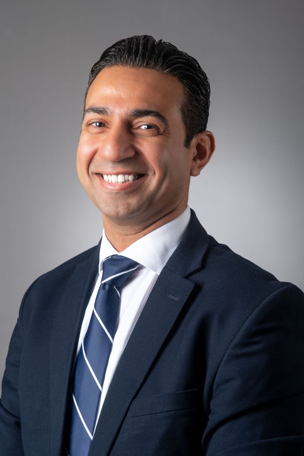 Professional headshot of a person in a navy suit and striped tie against a grey background.