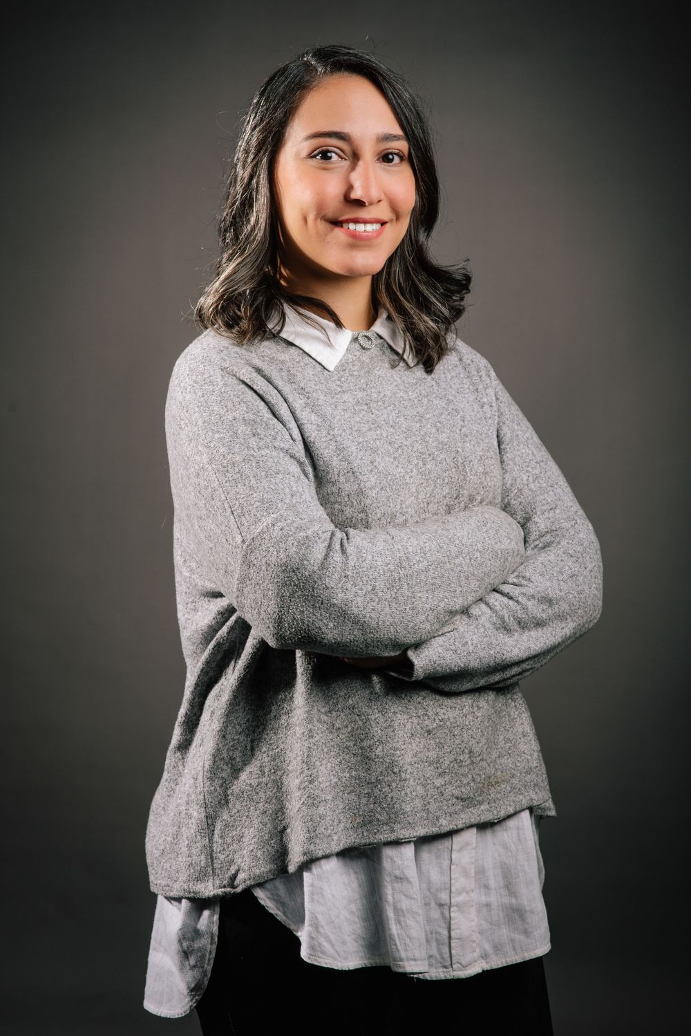 Professional portrait of person in layered gray sweater over white shirt against dark background.