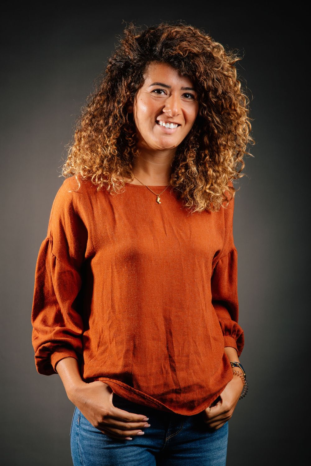 Casual portrait of person wearing burnt orange sweater with curly hair against dark backdrop.
