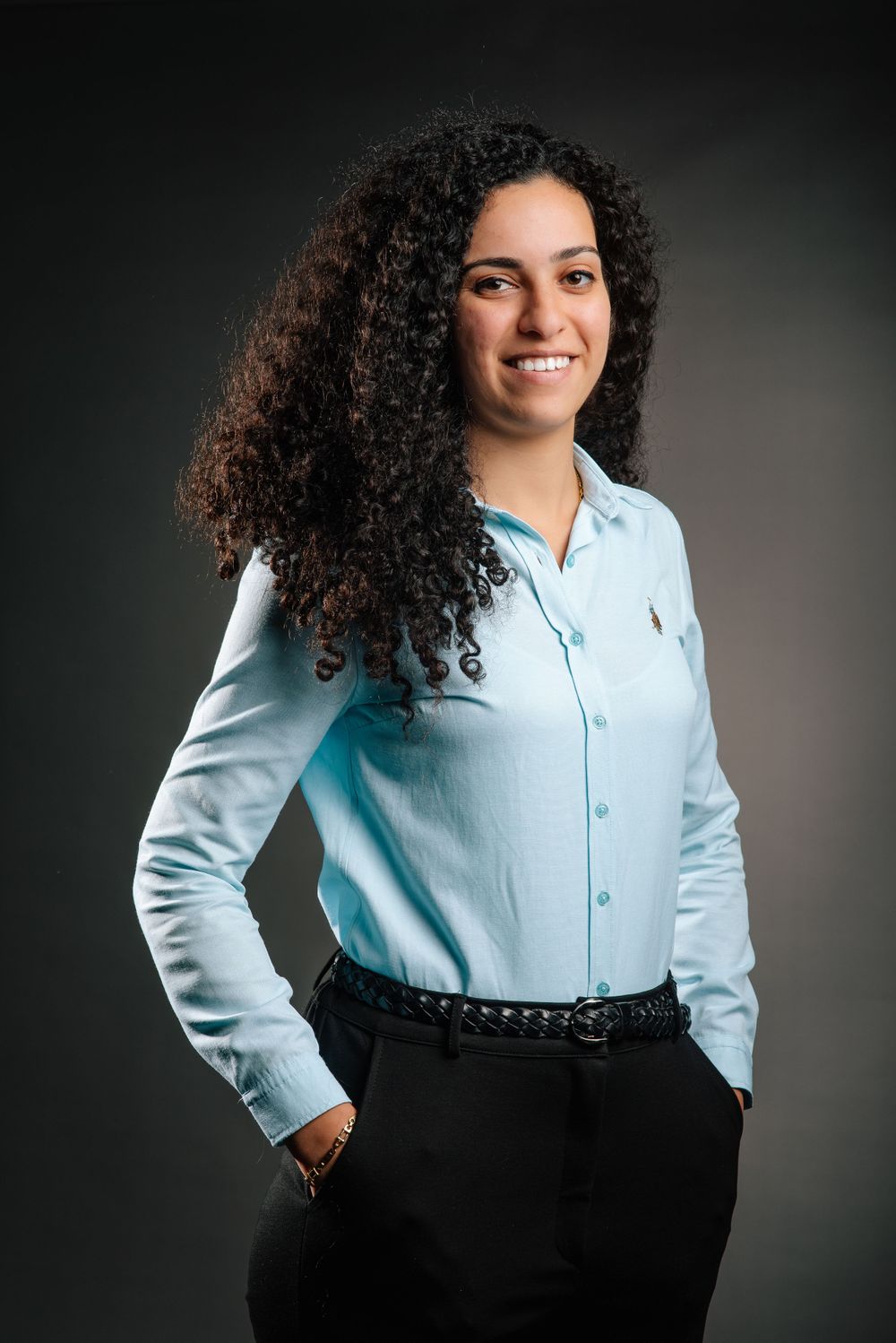 Professional portrait of a person in a light blue button-down shirt with natural curly hair against a dark background.