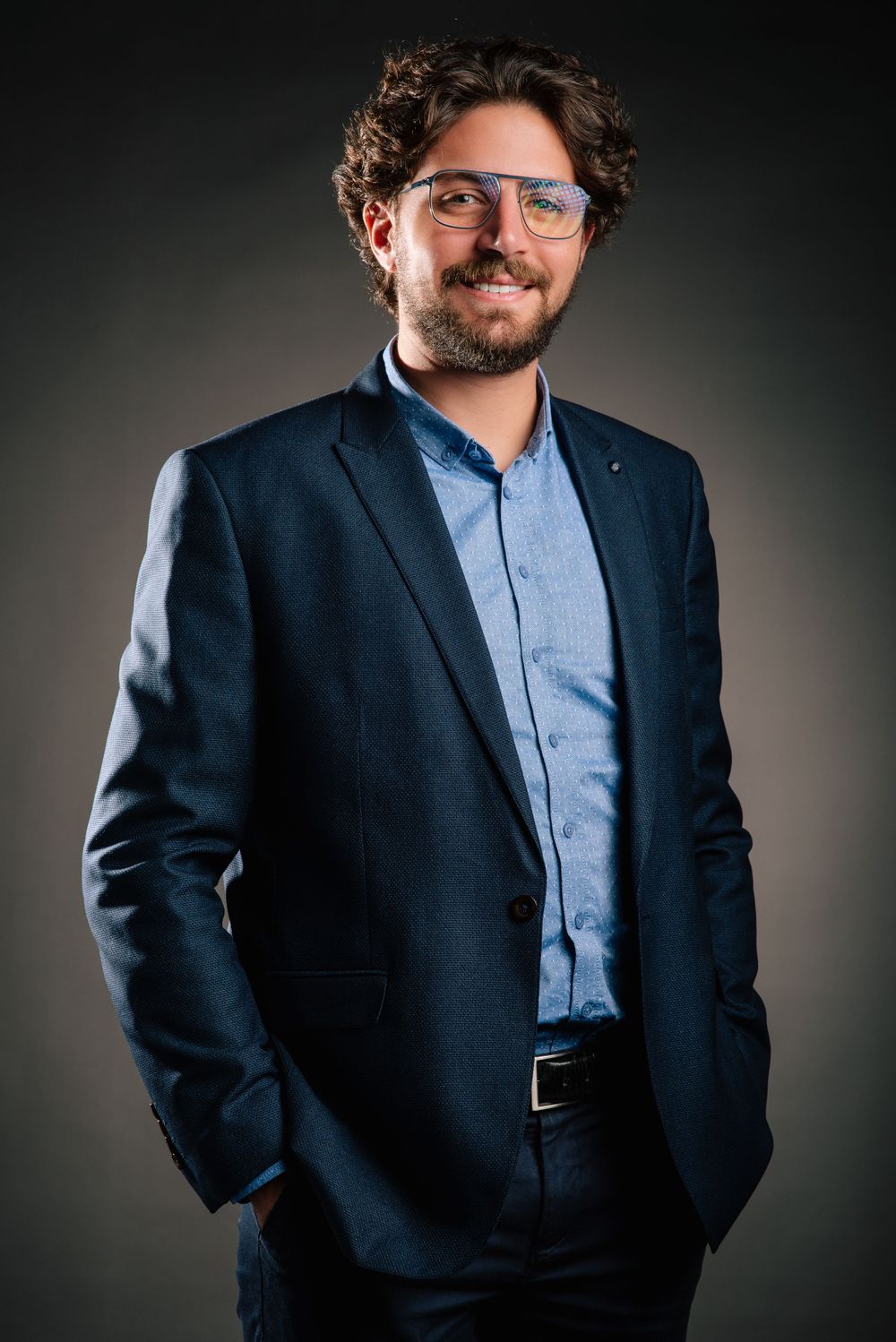 Professional wearing a navy suit and light blue shirt poses confidently against dark background with glasses.