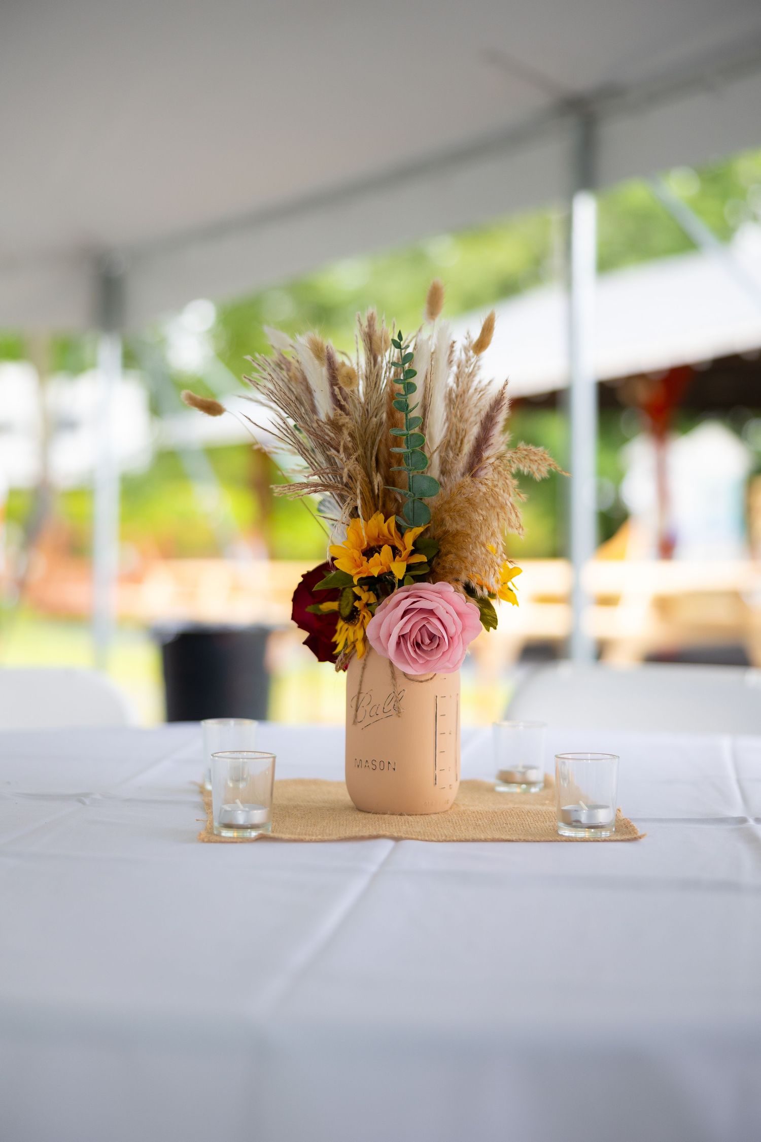 Mason jar centerpieces with sunflowers and pink roses on white tablecloths create an elegant rustic wedding setting.