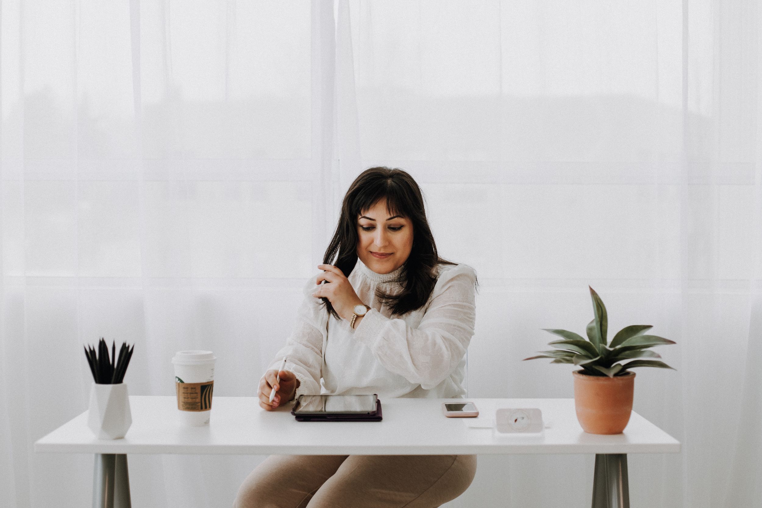Woman in white sweater working at minimalist desk with coffee and plant.