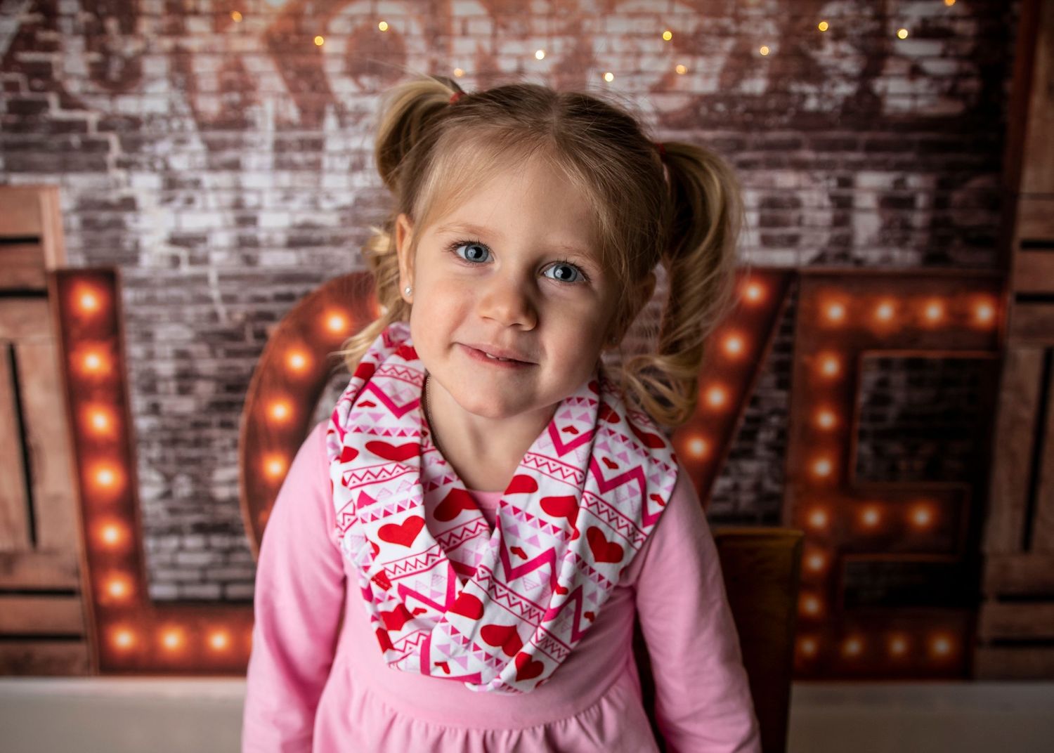 A child wearing pink with a heart-patterned scarf smiles in front of a brick wall with glowing letters.