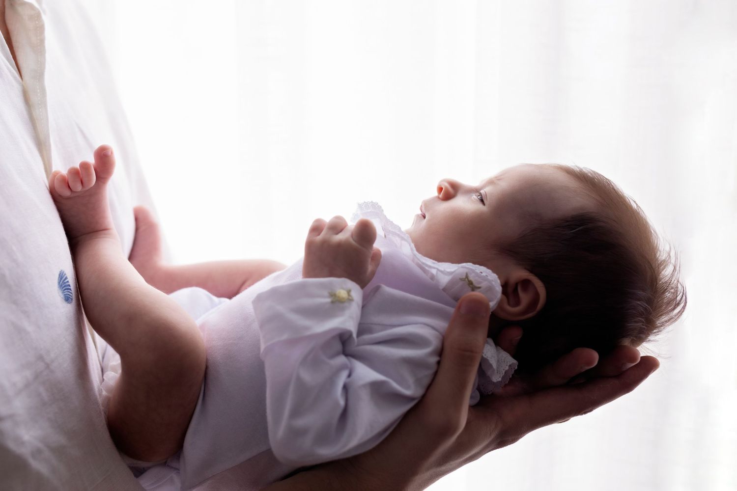 Delicate portrait of baby in white onesie being cradled against bright background.
