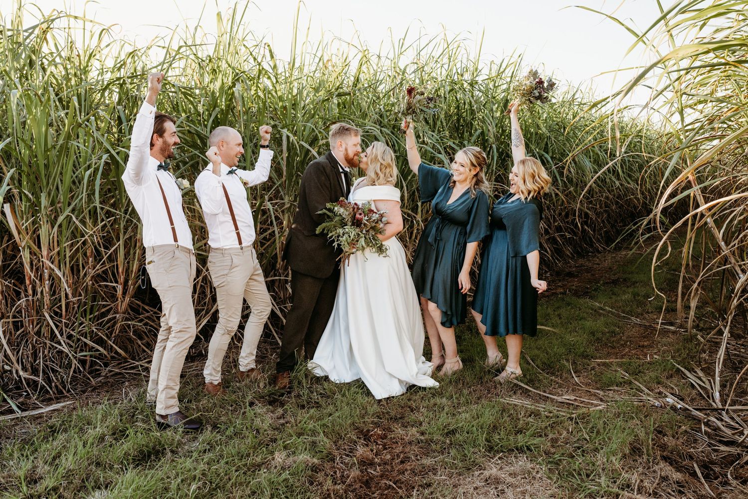 Wedding party jumping and celebrating in front of tall grass outdoors.