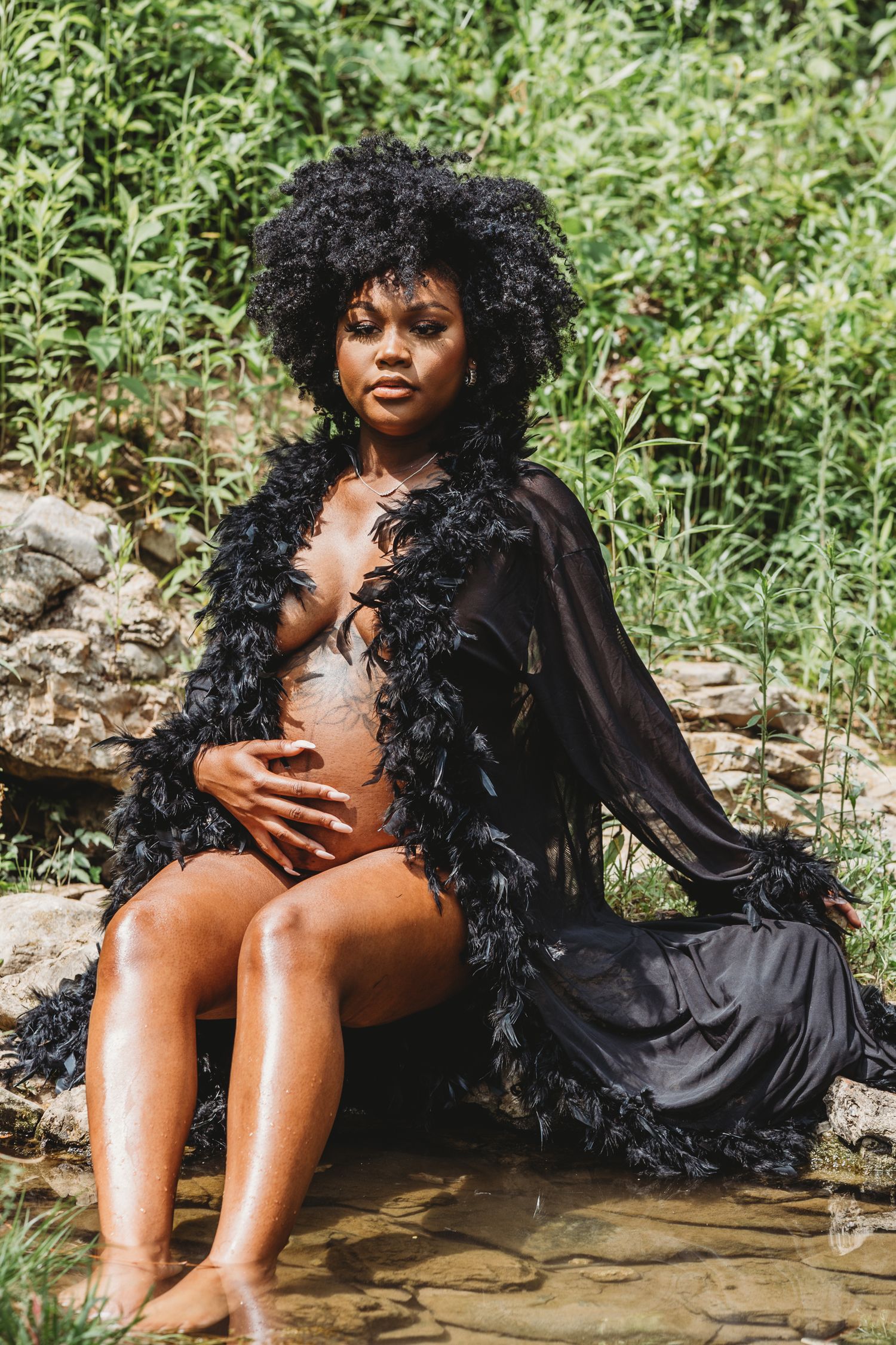 Figure in a flowing black feathered robe poses artistically while sitting on rocks near water in a natural setting.