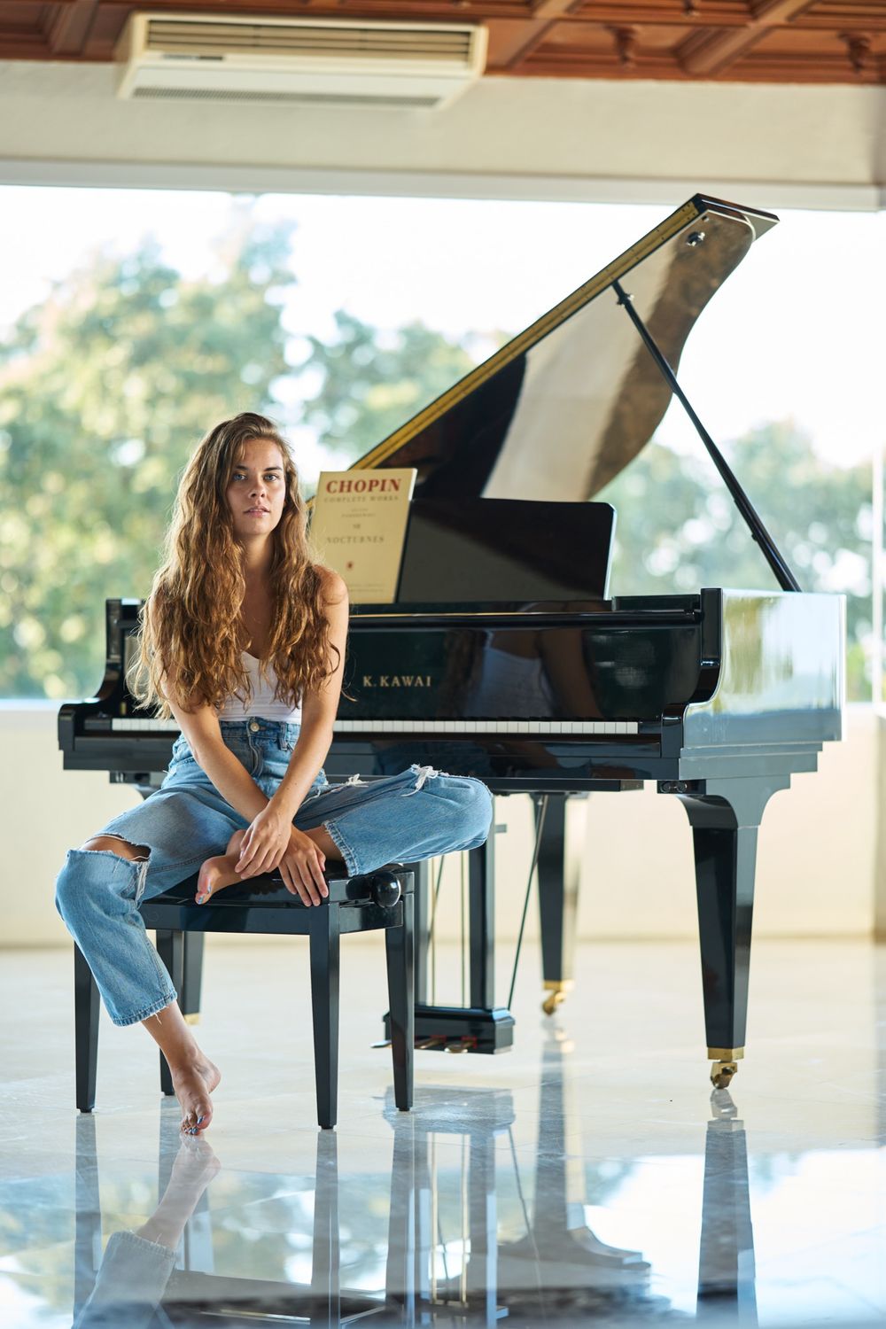 Casual portrait of a pianist sitting on a grand piano bench in a sunlit room.