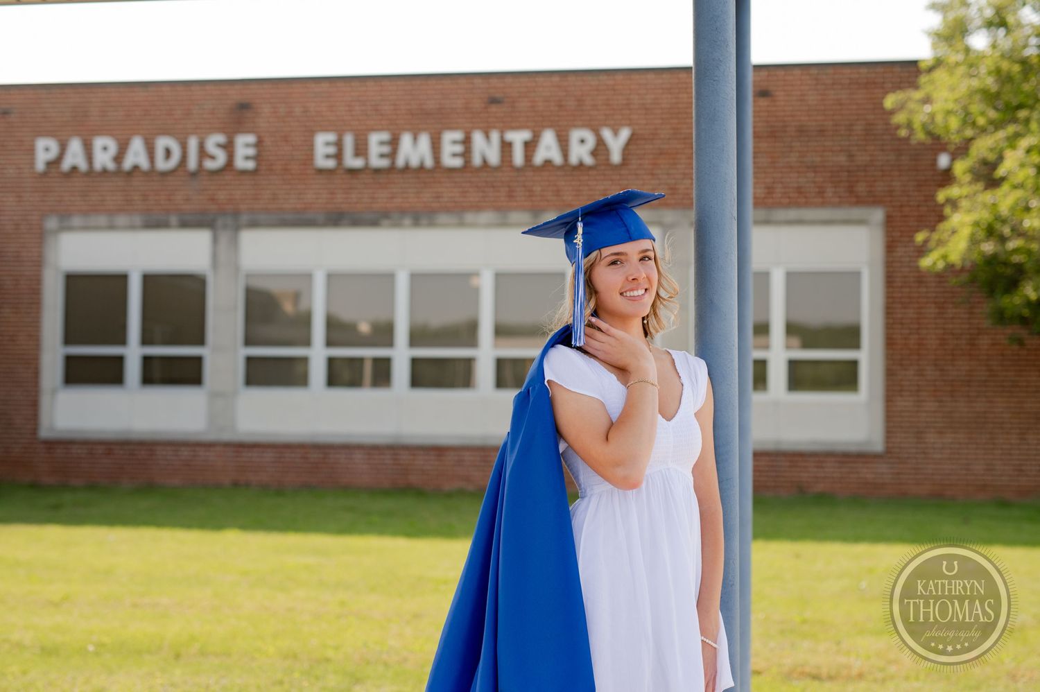 Graduate in white dress and blue cap and gown poses by support column at elementary school.