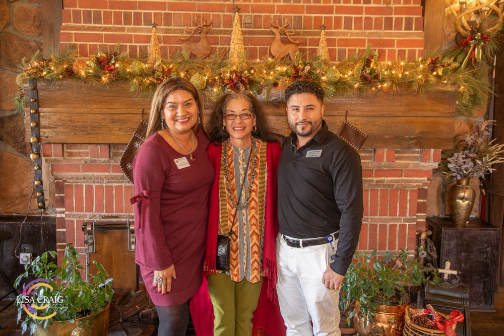 Three people pose by a decorated brick fireplace mantel with holiday greenery and decor.