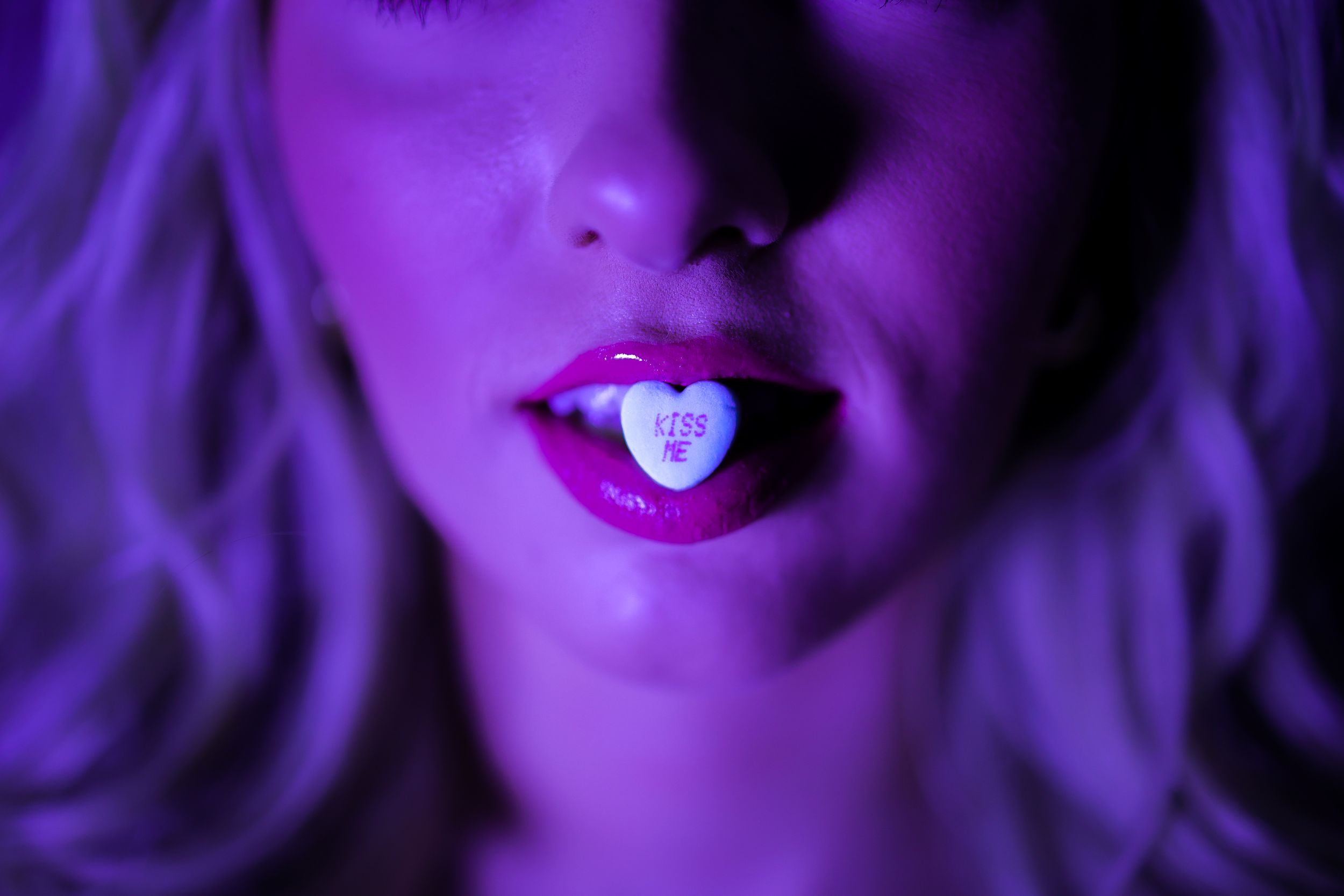 Close-up of lips holding a small heart-shaped candy in purple neon lighting.