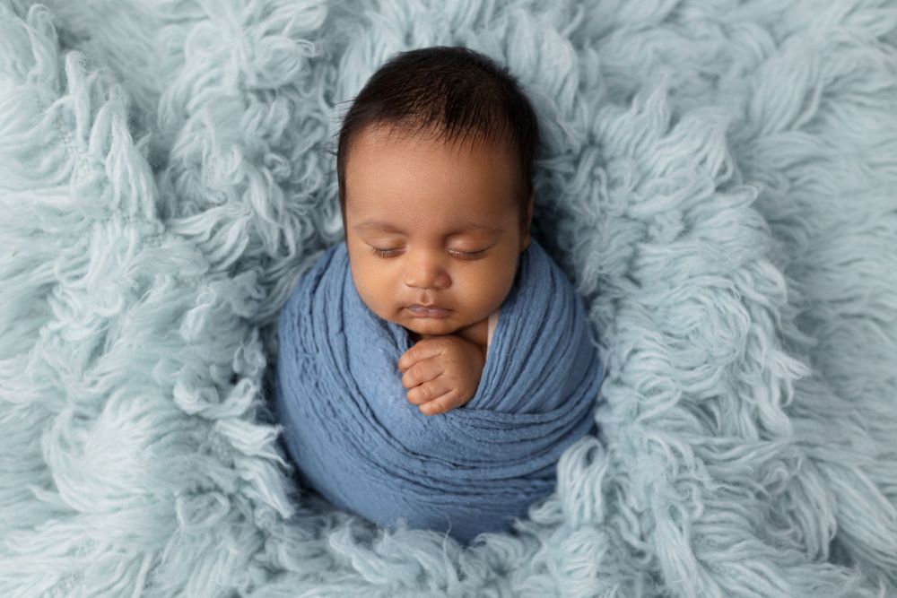 Peaceful newborn wrapped in soft blue fabric on fuzzy teal blanket.
