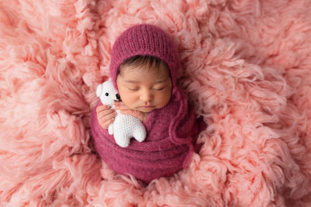 Sleeping newborn in burgundy wrap with knit hat and toy on pink fur background.