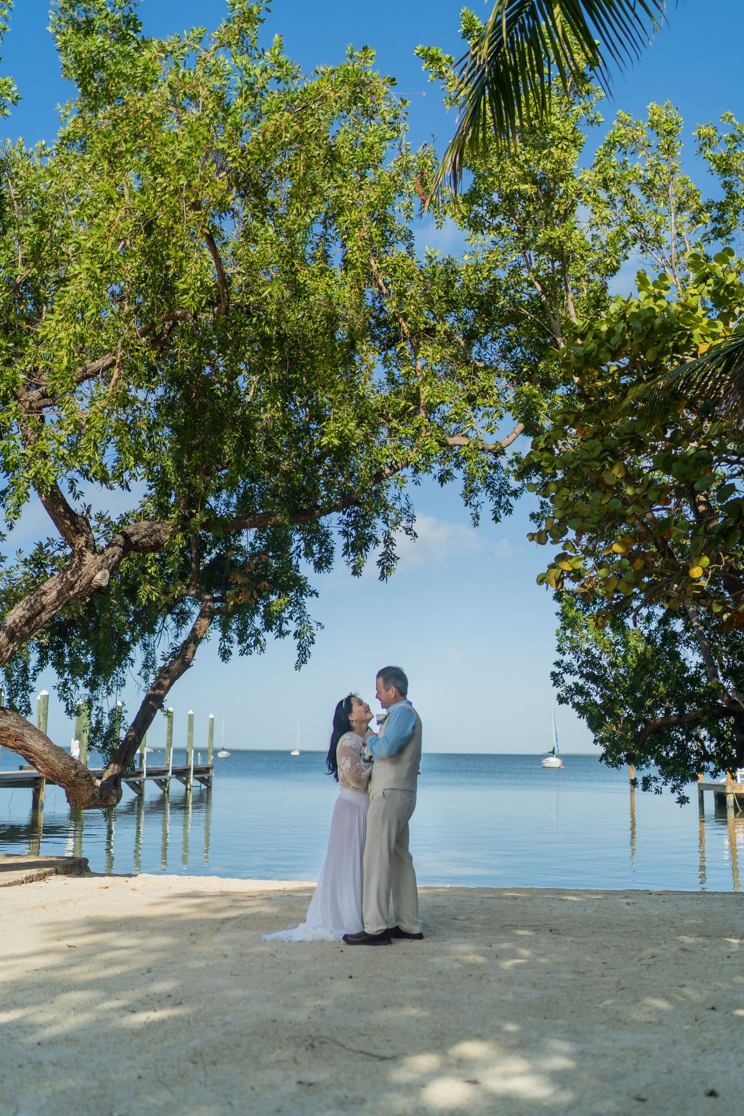 Couple shares intimate moment under trees by oceanfront dock during tropical wedding ceremony.