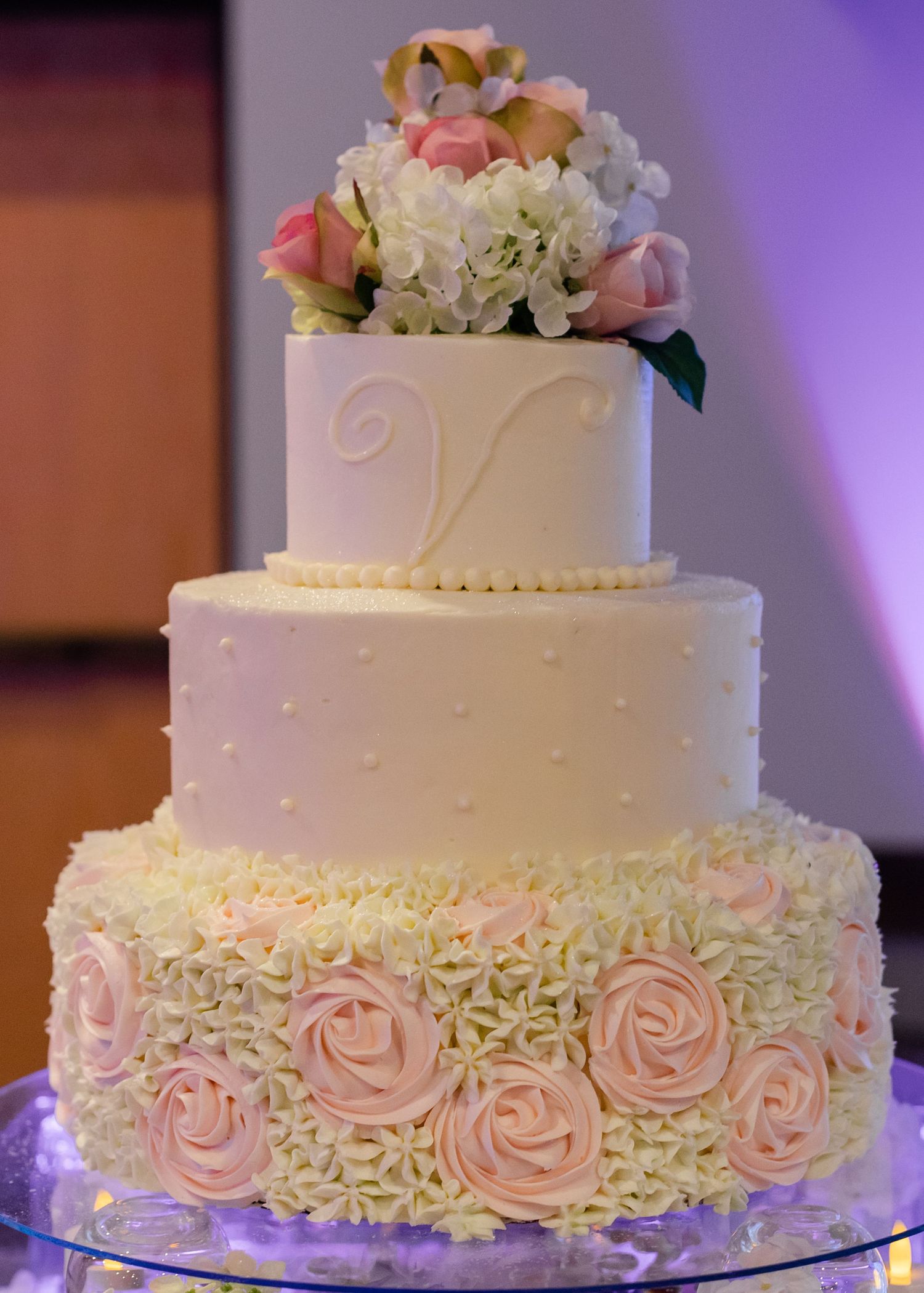 Elegant white wedding cake with delicate pink roses and intricate lace piping details displayed at a reception.
