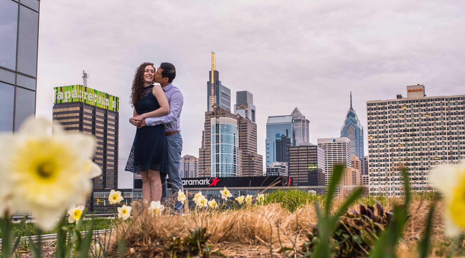 Urban couple embraces near blooming yellow flowers with Philadelphia's downtown towers in background.