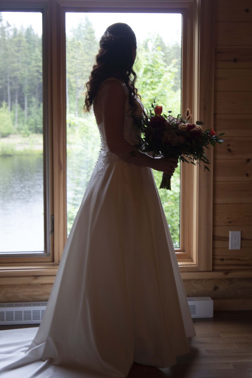 A silhouette of a bride in a white wedding gown standing by a window overlooking a lake and forest.