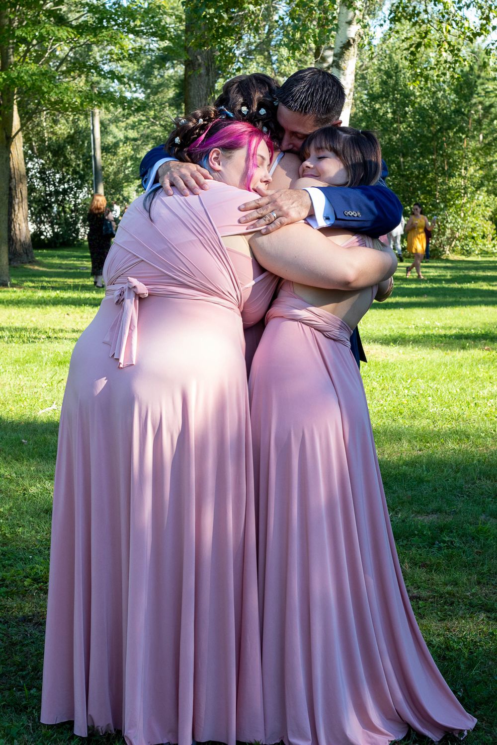Three bridesmaids in pink dresses sharing an emotional group hug outdoors.