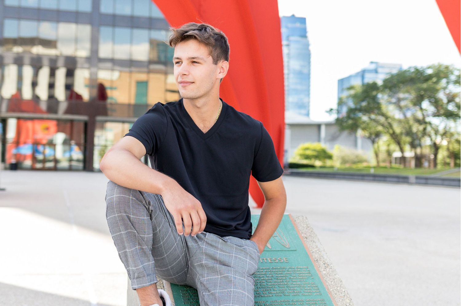 Young man in black t-shirt and plaid pants sitting outdoors in an urban setting.