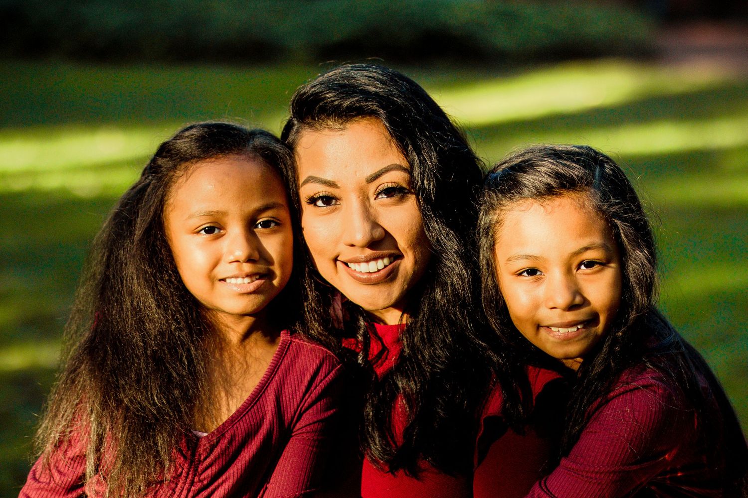 A smiling mother poses outdoors with her two young daughters wearing matching red outfits.