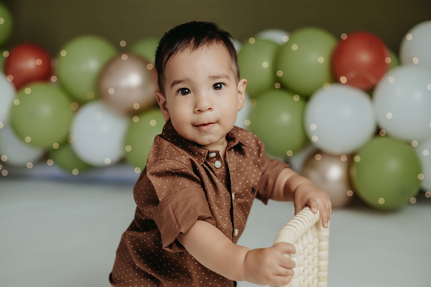 Young child in brown shirt poses against festive balloon backdrop at party.