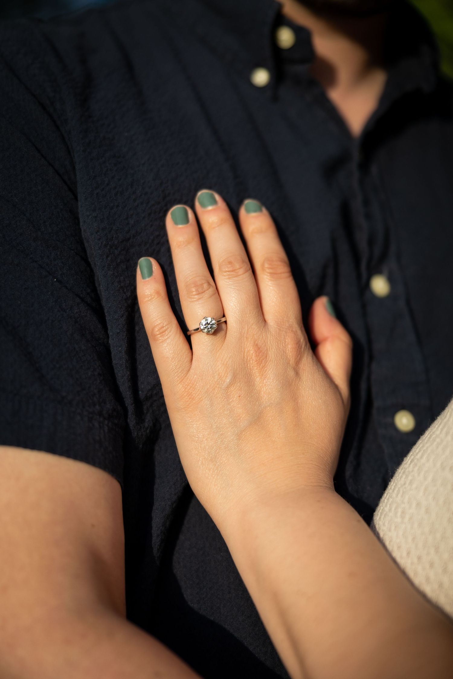 Close-up of a hand with teal nail polish wearing an engagement ring against a black shirt.