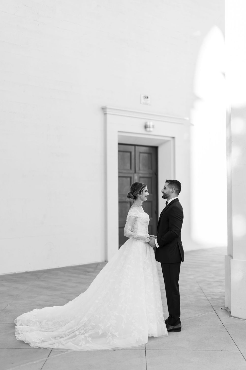 Black and white portrait of wedding couple standing before wooden door in classic attire.