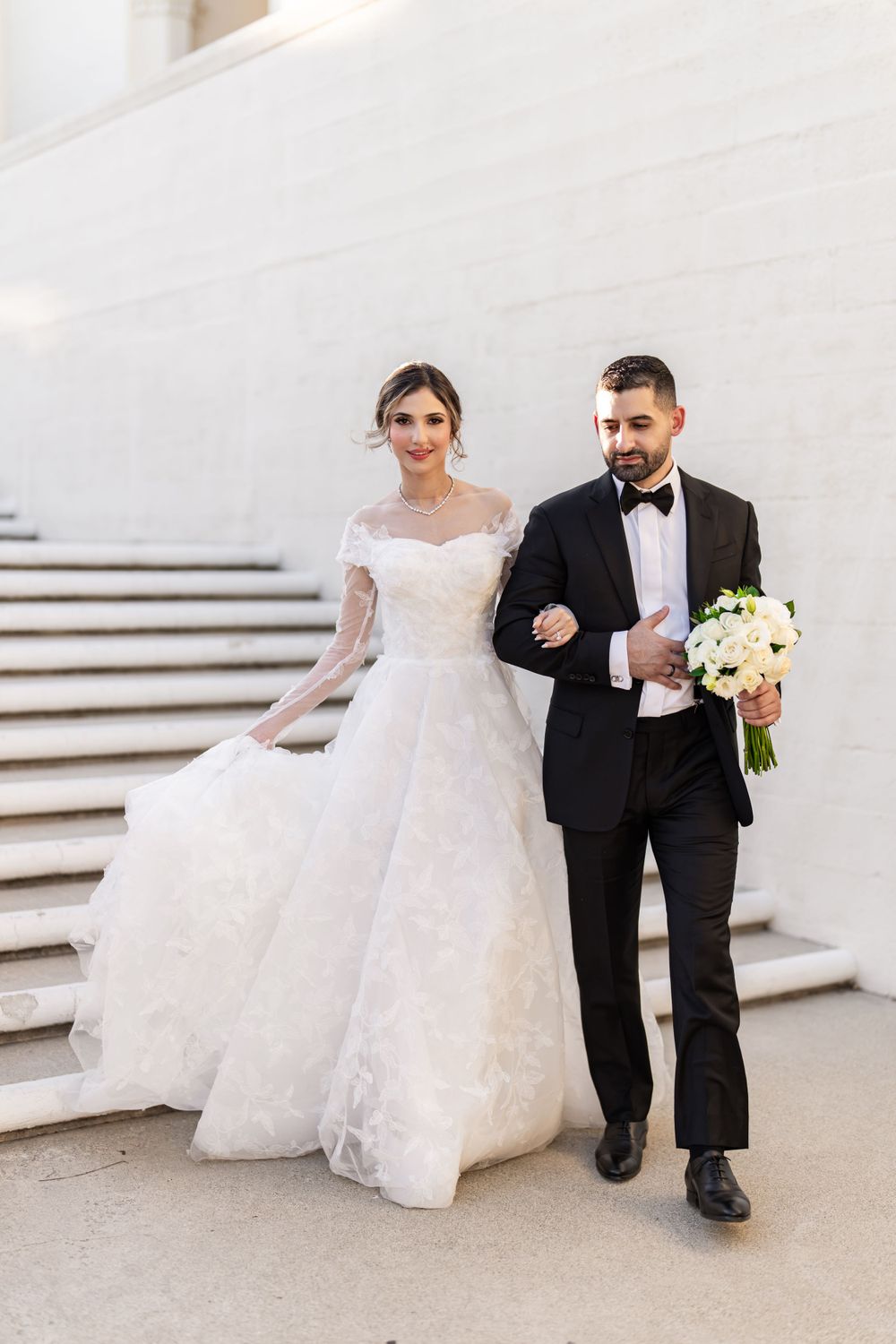 Wedding couple descending white stairs, bride in lace gown and groom in classic tuxedo.