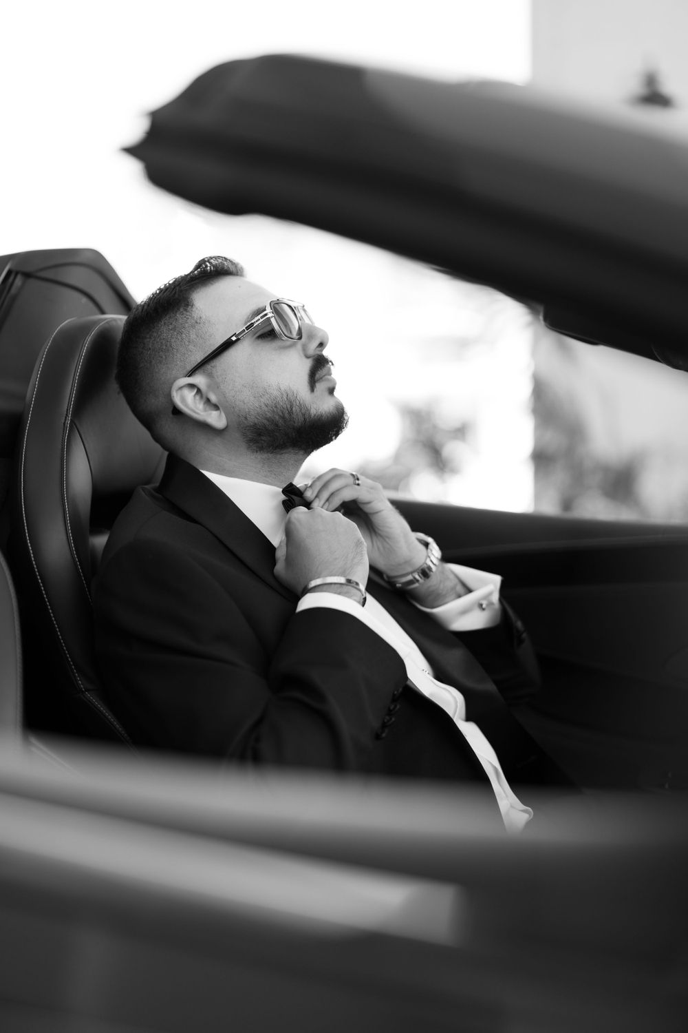 Groom in black suit adjusts tie while seated in luxury car.