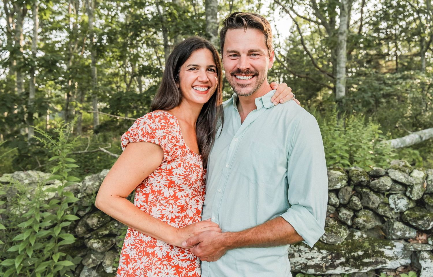 Couple in floral dress and light blue shirt pose together against greenery and stone wall.
