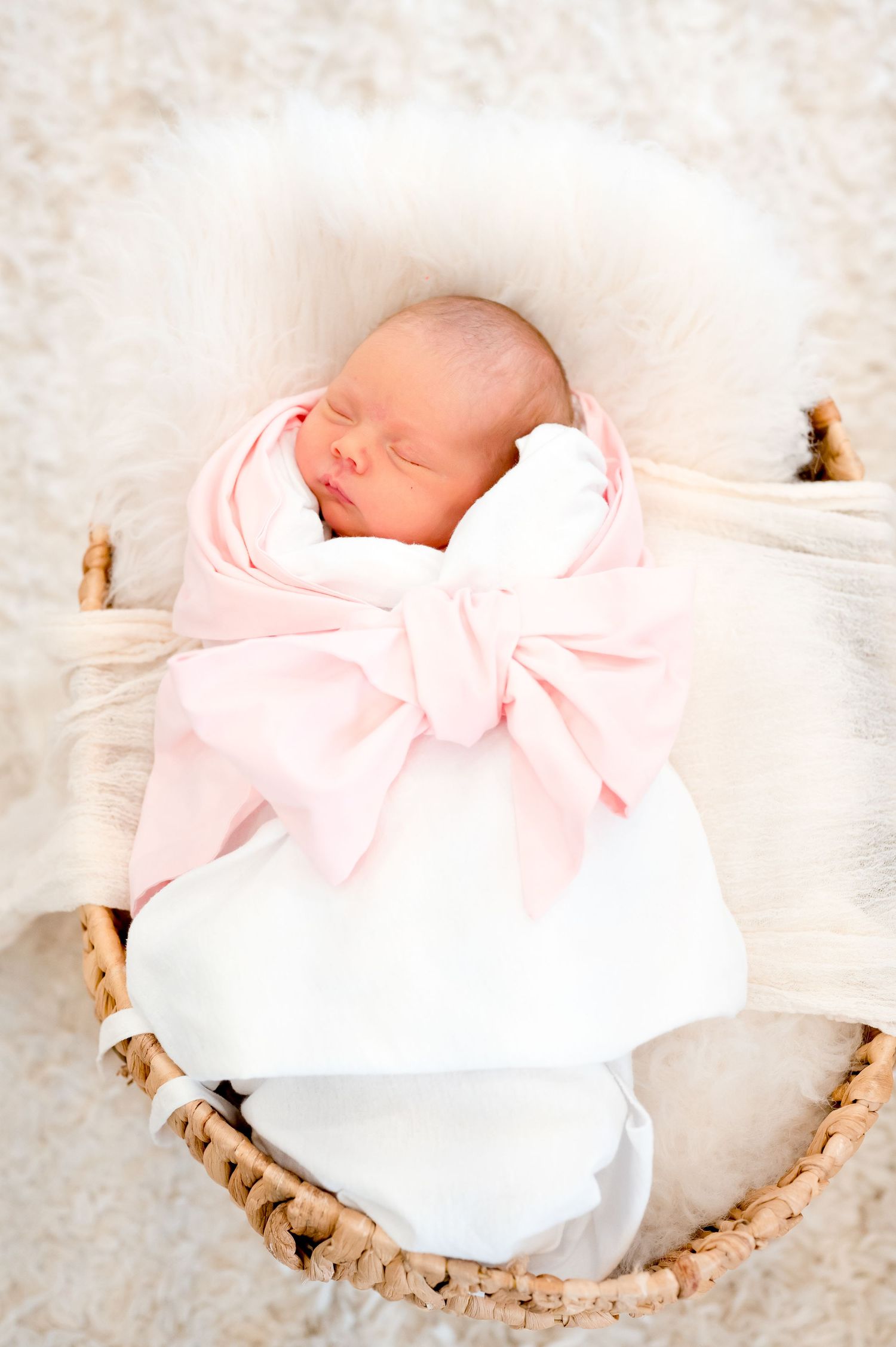 light and bright photo of a newborn baby girl in a basket wearing a white wrap and pink bow