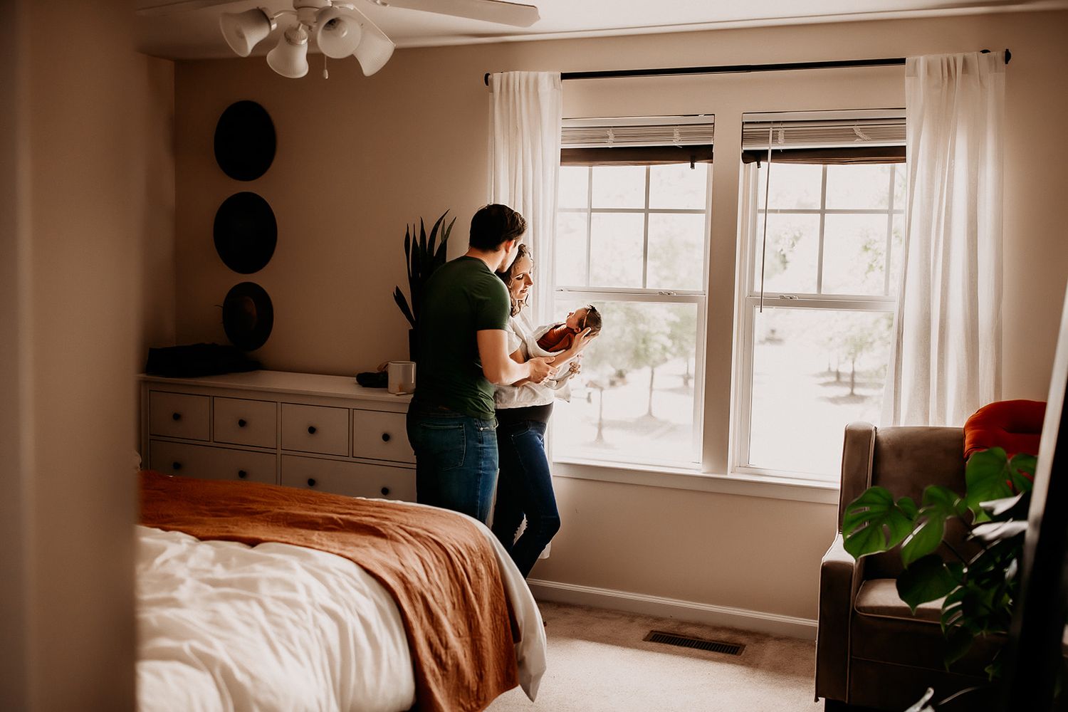 A mother and father stand in front of the window in their master bedroom looking down at their newborn baby girl.